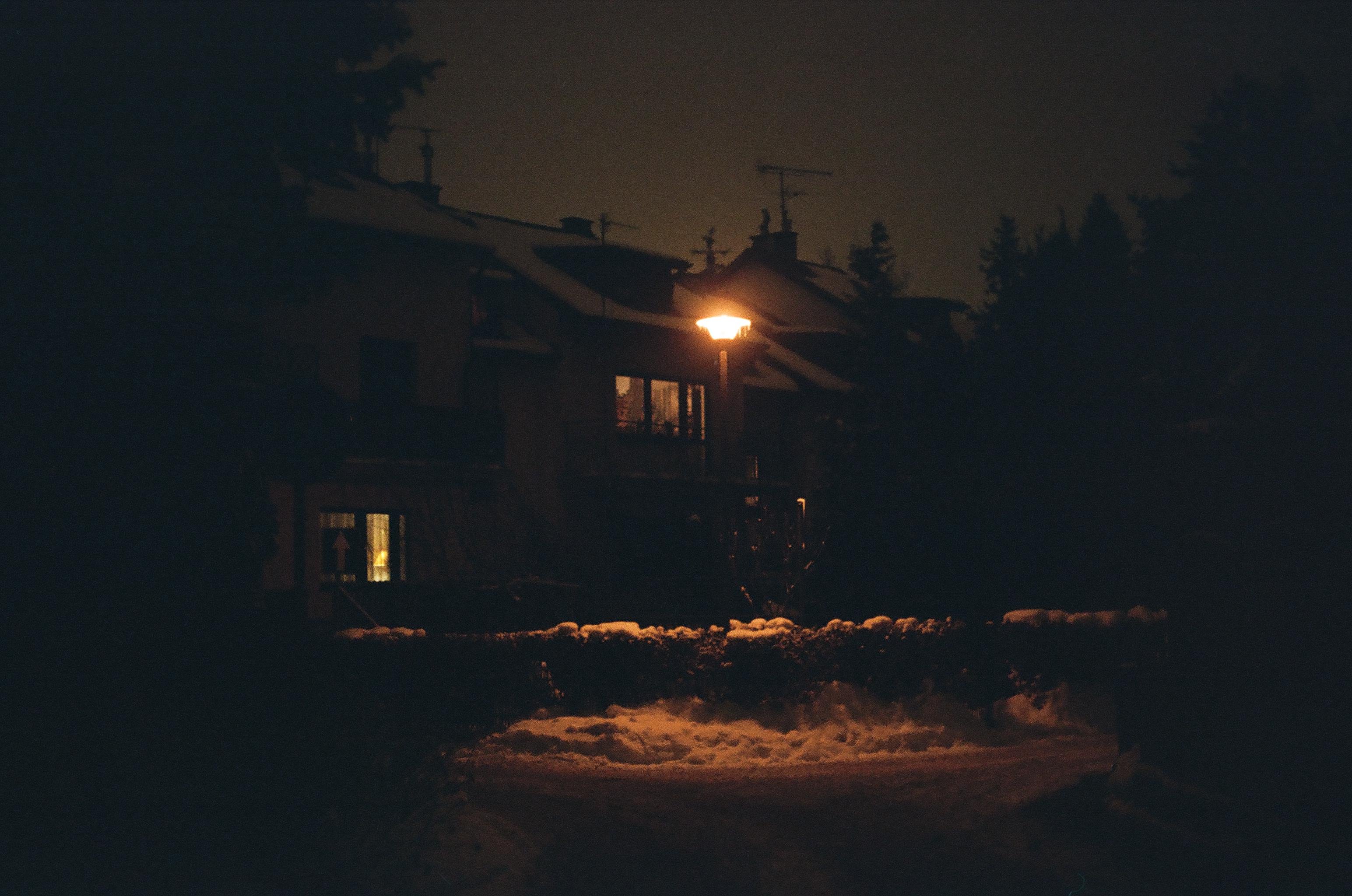 Dimly lit apartment buildings at night with snow on the ground, a glowing streetlamp near the center, and a few illuminated windows, framed by dark blurred edges.