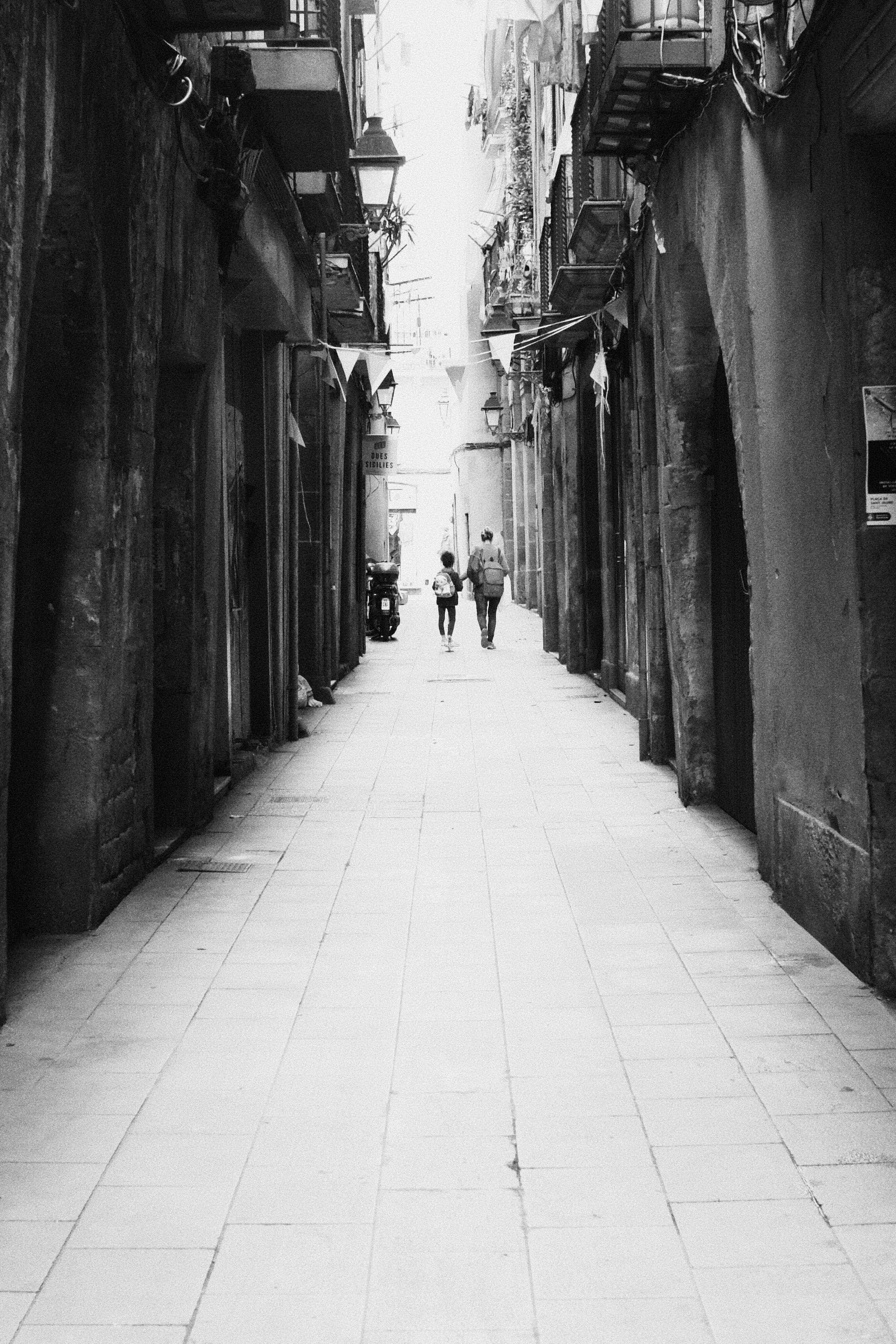 Schwarz-Weiß-Foto einer engen Gasse mit hohen Gebäuden und Balkonen auf beiden Seiten, hängende Straßenlaternen und Fahnen über dem Kopf, und zwei Menschen, die zu einer entlang der Gasse gehen; ein Roller ist entlang der linken Wand geparkt.