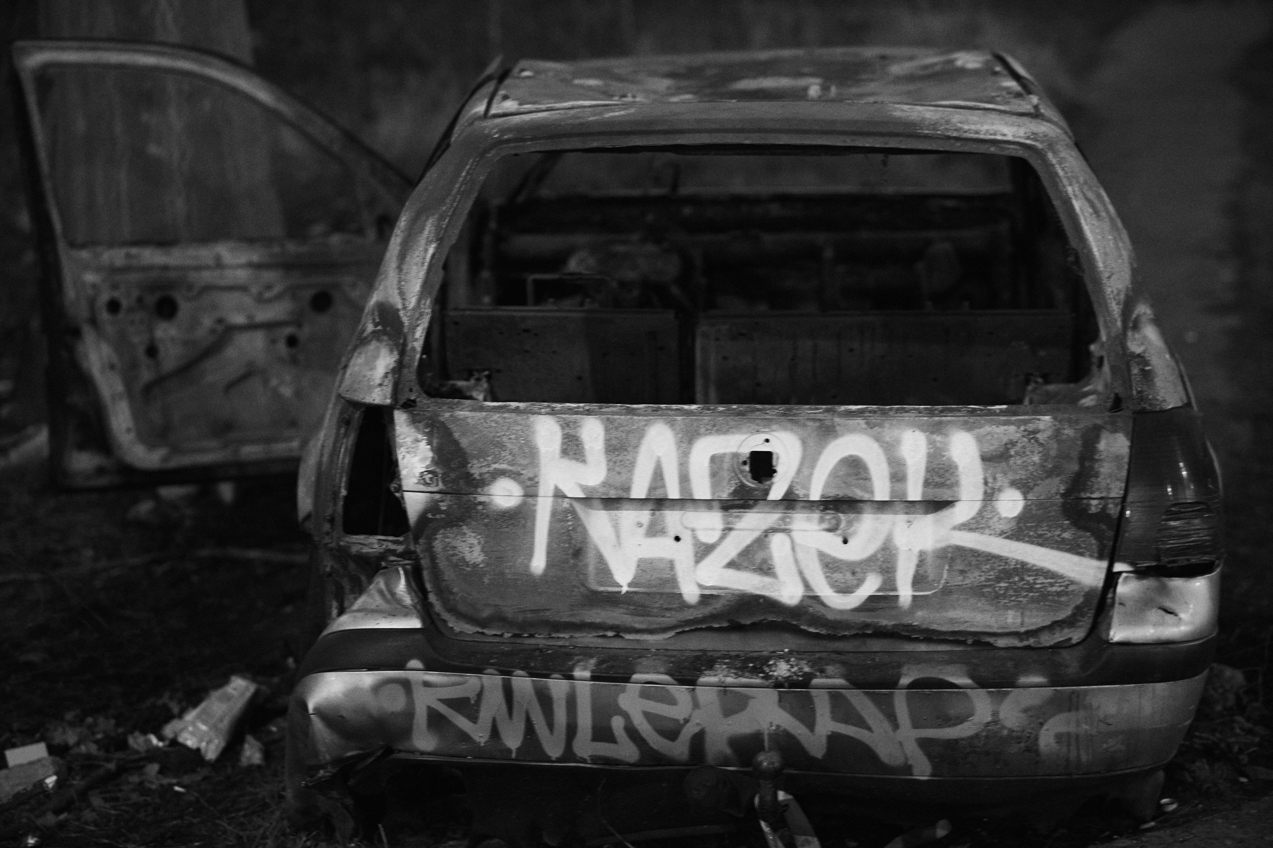 The image shows a burned-out car with its door open. The exterior is covered in graffiti. The setting appears to be outdoors, and the car looks abandoned.