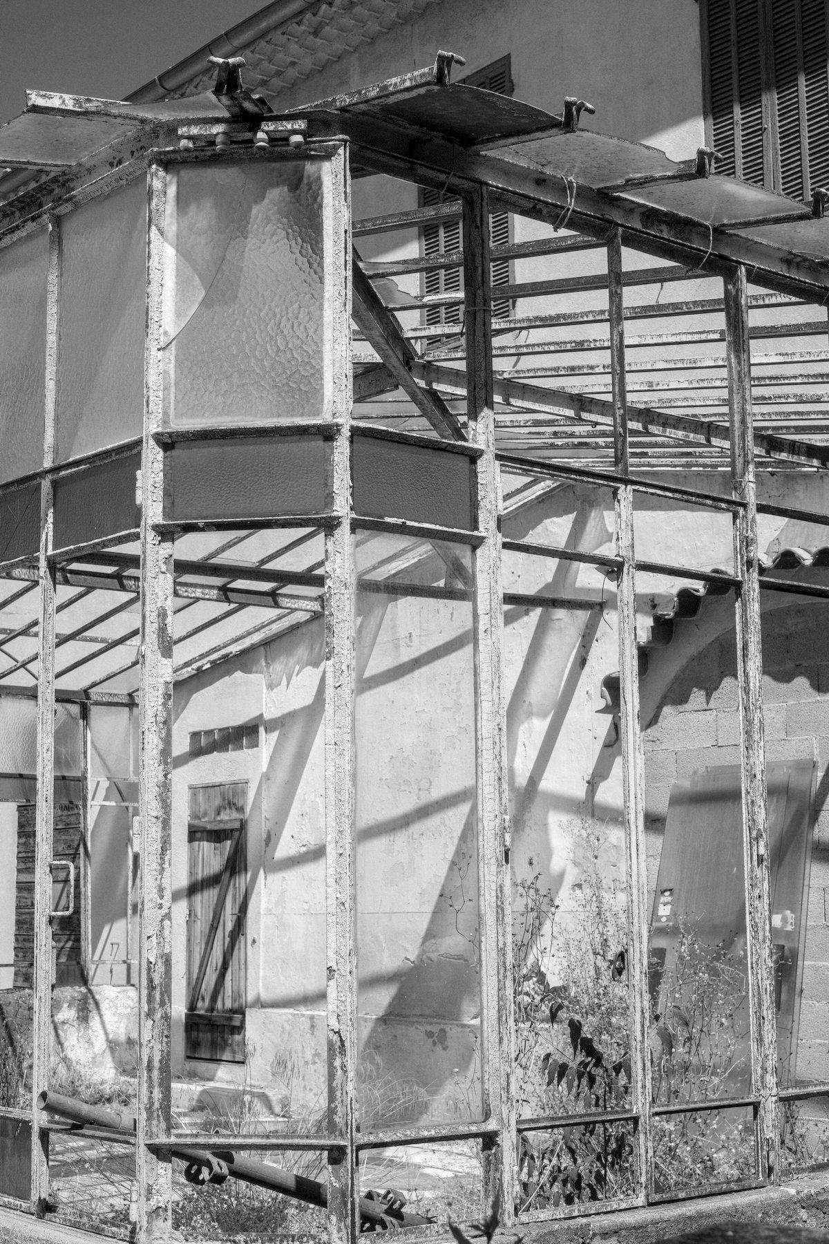 Photo verticale en noir et blanc. Au premier plan, un coin de véranda avec la majorité des vitres manquantes. À l’intérieur, à droite, 2 portes en verre appuyées contre le mur de la bâtisse. Au fond à gauche de la photo, une porte en bois fermée