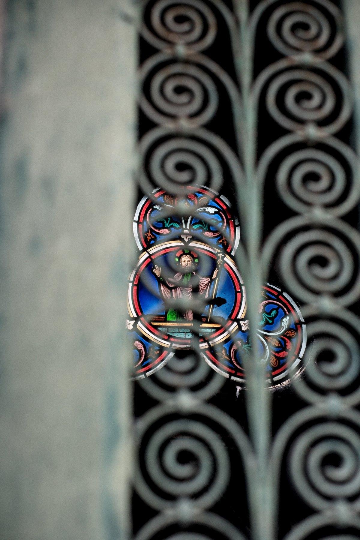 Photo verticale. Gros plan sur une porte de mausolée avec des spirales en fer forgé, à travers l’une d’elles, un vitrail sur le mur du fond, la tête de Jésus au centre d’une spirale