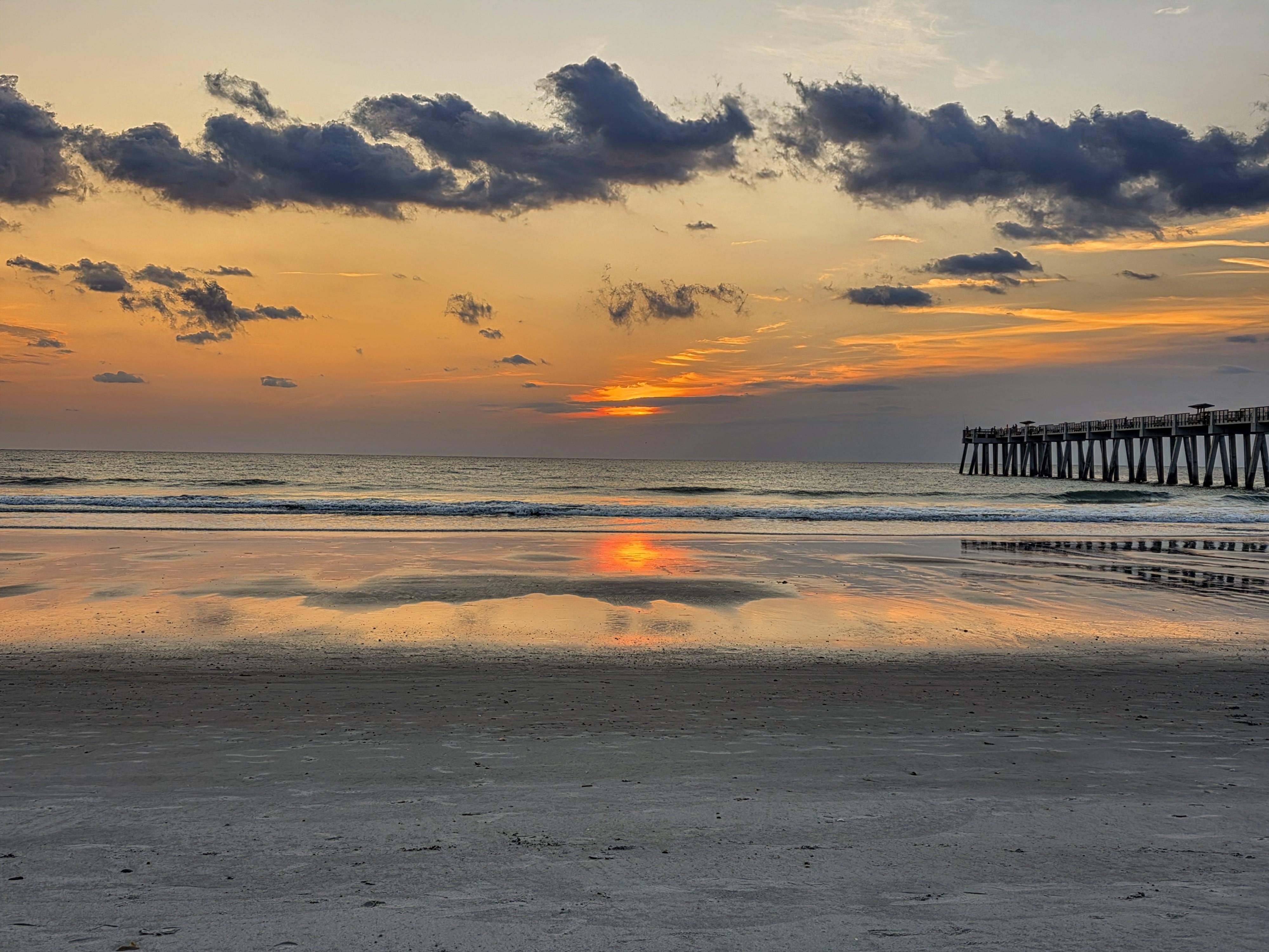 Surise over a sandy beach with orange and yellow hues lighting the sky. Dark clouds are scattered across the horizon. A long pier extends from the right side into the ocean. The wet sand reflects the colors of the sky and rising sun. Waves gently reach the shore.