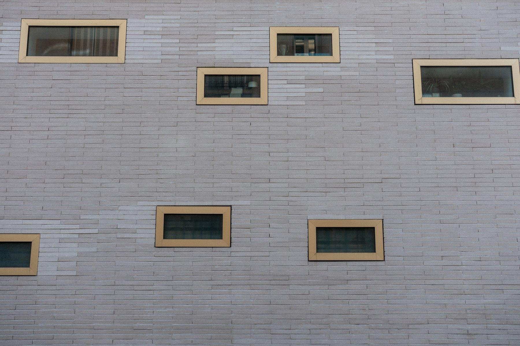 Photo horizontale. Un mur fait de petites dalles grises rectangulaires assez longues, horizontales, dans lequel des petite fenêtres rectangulaires horizontales, au cadre jaune, de tailles différentes et placées sans logique apparente