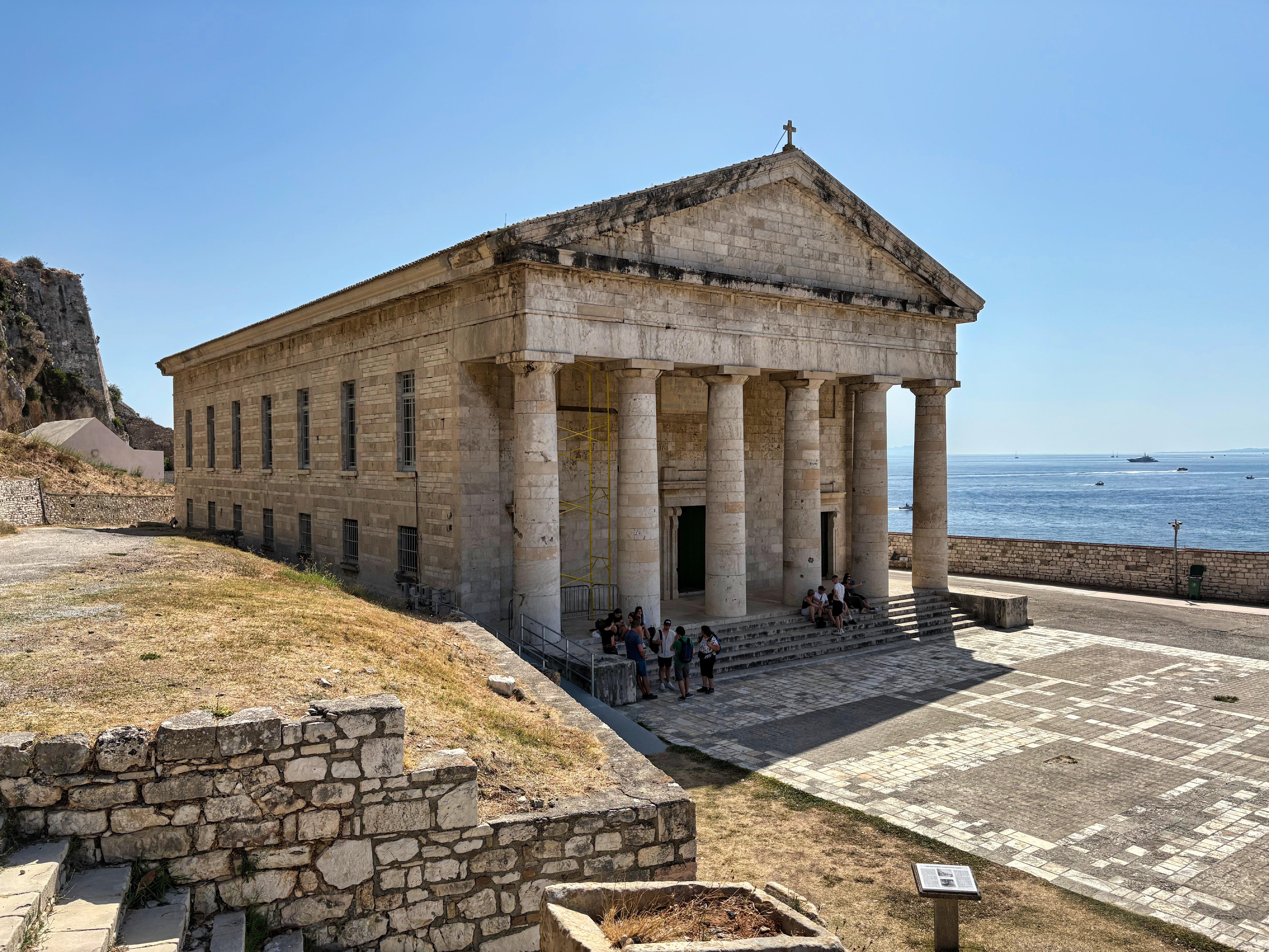 Stone building with classical Greek architecture, featuring large columns and a triangular pediment, located near the sea. A group of people is gathered at the entrance, sitting on steps under the portico. Stone wall and pathway in the foreground, with grassy area on the left leading to the corner of the building. Water and clear sky in the background.