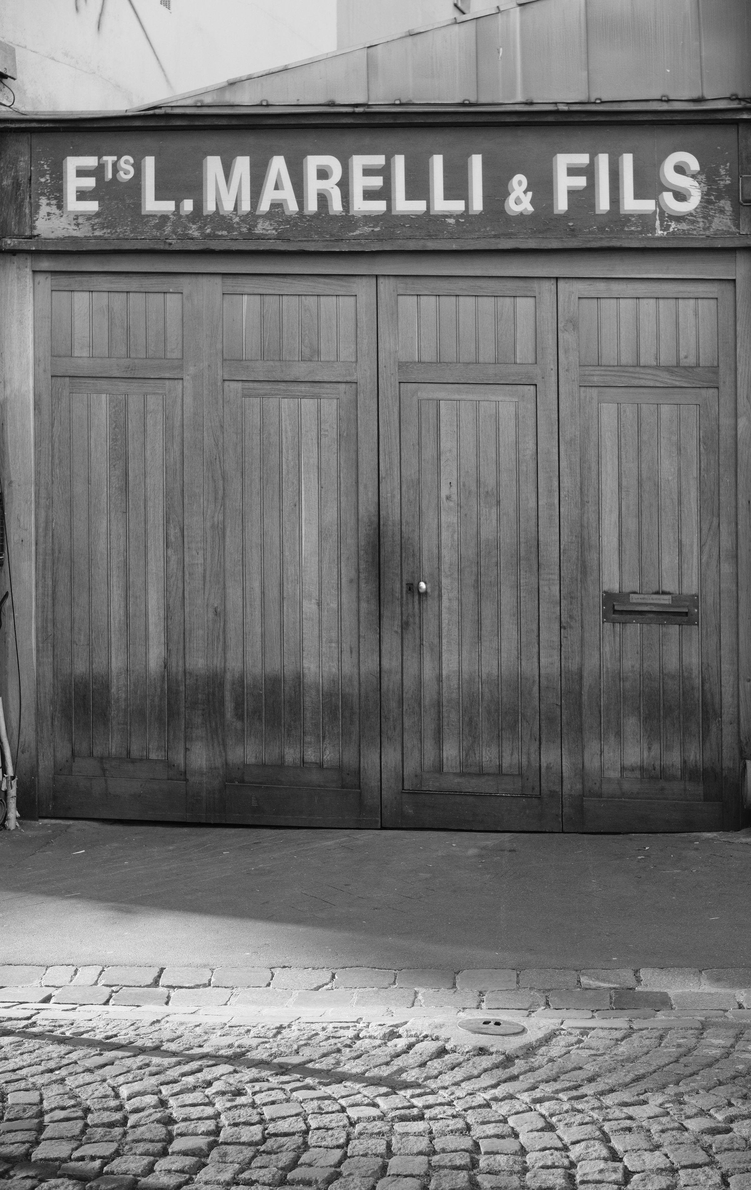 Photo format portrait en noir et blanc d’une façade de commerce en bois, avec l’enseigne en haut disant « Ets L. Marelli & Fils ». Le sol est pavé en arc de cercles au premier plan et éclairé par des rayons du soleil venant de la gauche de l’image