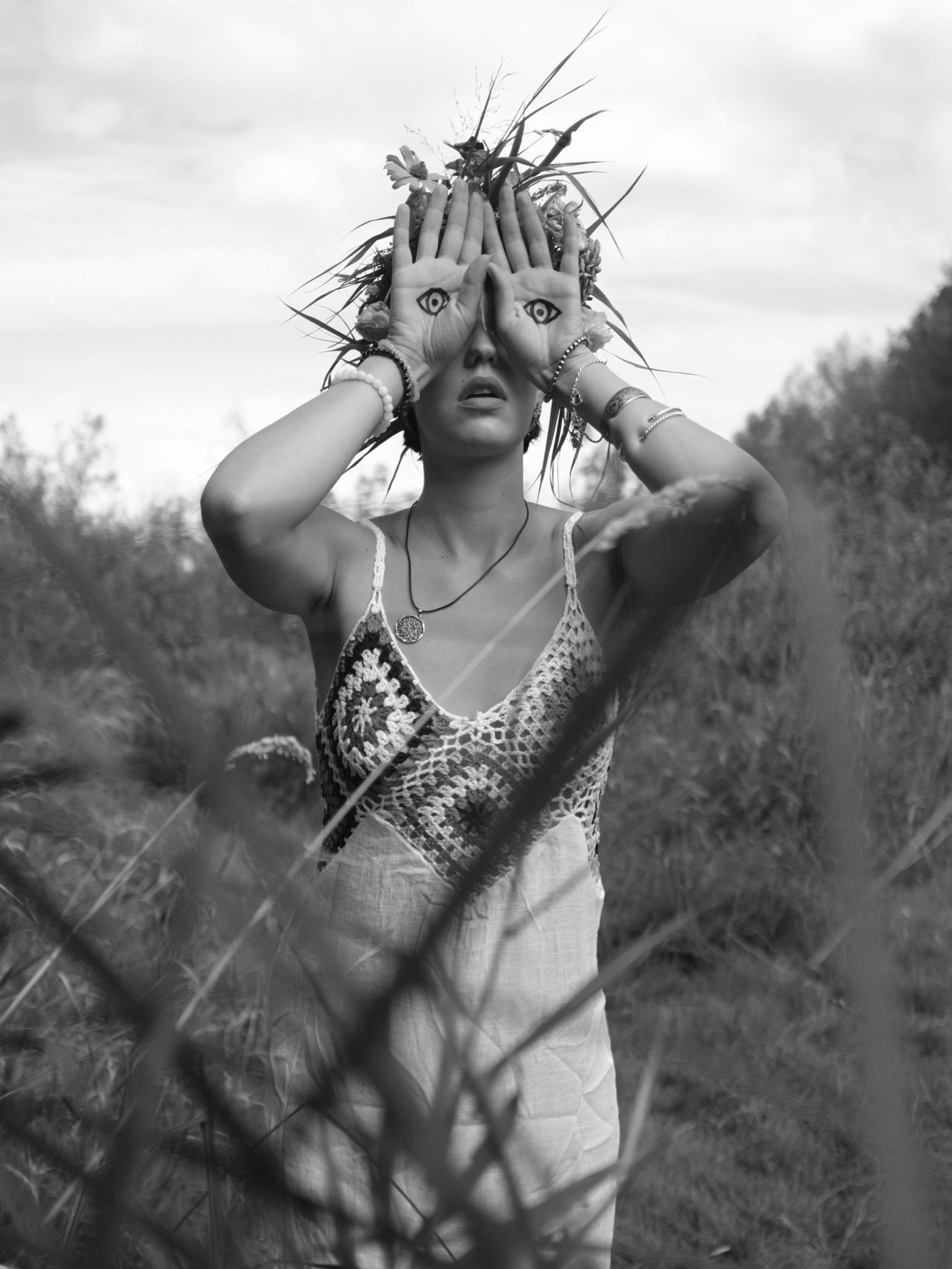 Person standing outdoors in a grassy area, wearing a crocheted top and a headdress made of flowers and twigs. Hands are positioned over the face with eyes painted on the palms, creating an illusion of having eyes on the hands. The image is in black and white.