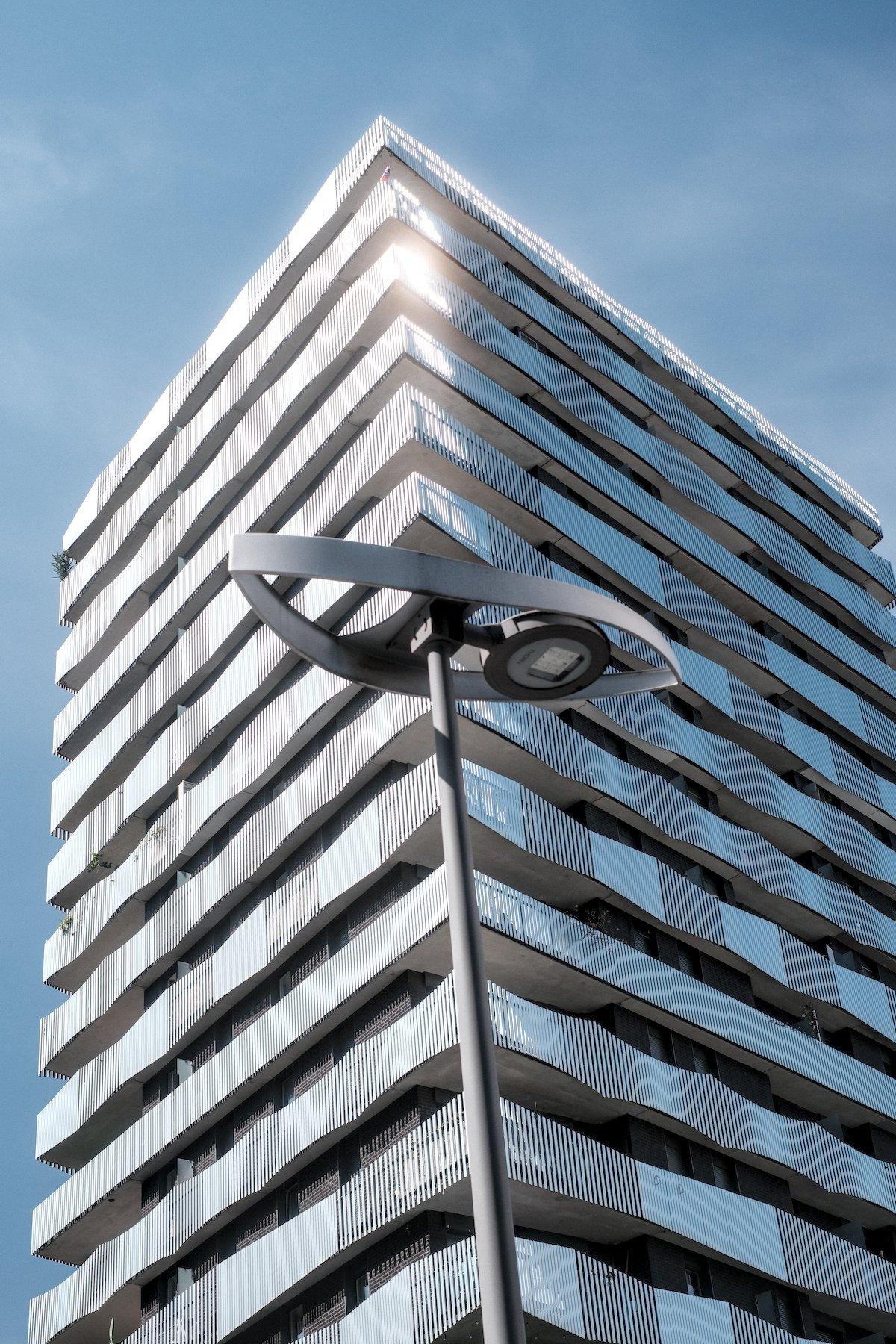 Photo verticale. Contre plongée sur un immeuble moderne, de forme traditionnelle mais avec des balcons aux 3 silhouettes se suivant d’un étage sur l’autre, mais tous avec une alternance plaque de métal pleine et surface pleine de fentes fines