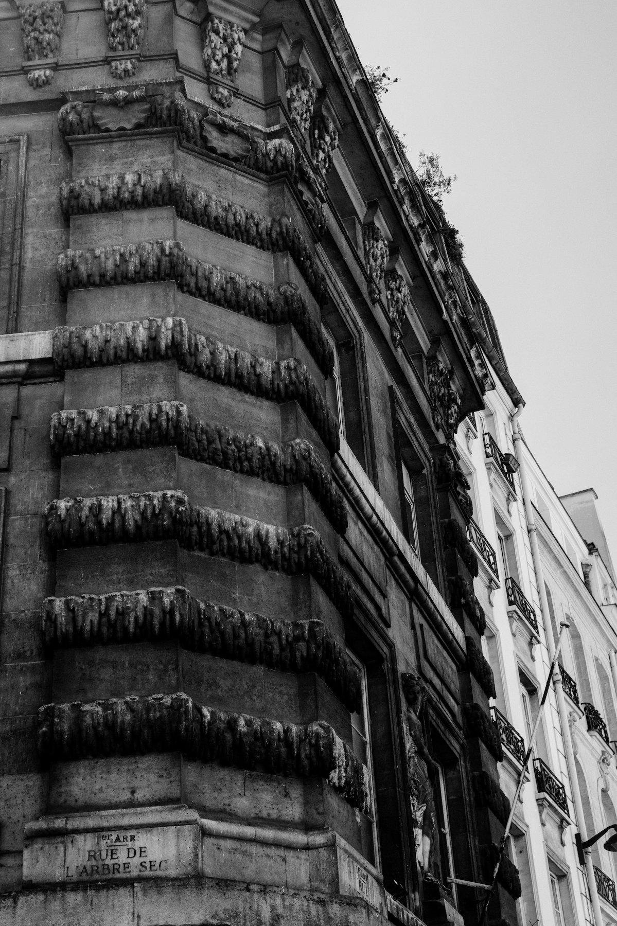 Photo verticale en noir et blanc. Un coin de bâtiment, à l’angle de la rue de l’arbre sec. Le nom de rue est gravée dans la pierre. L’angle du bâtiment présente des moulures en forme de feuilles de vigne