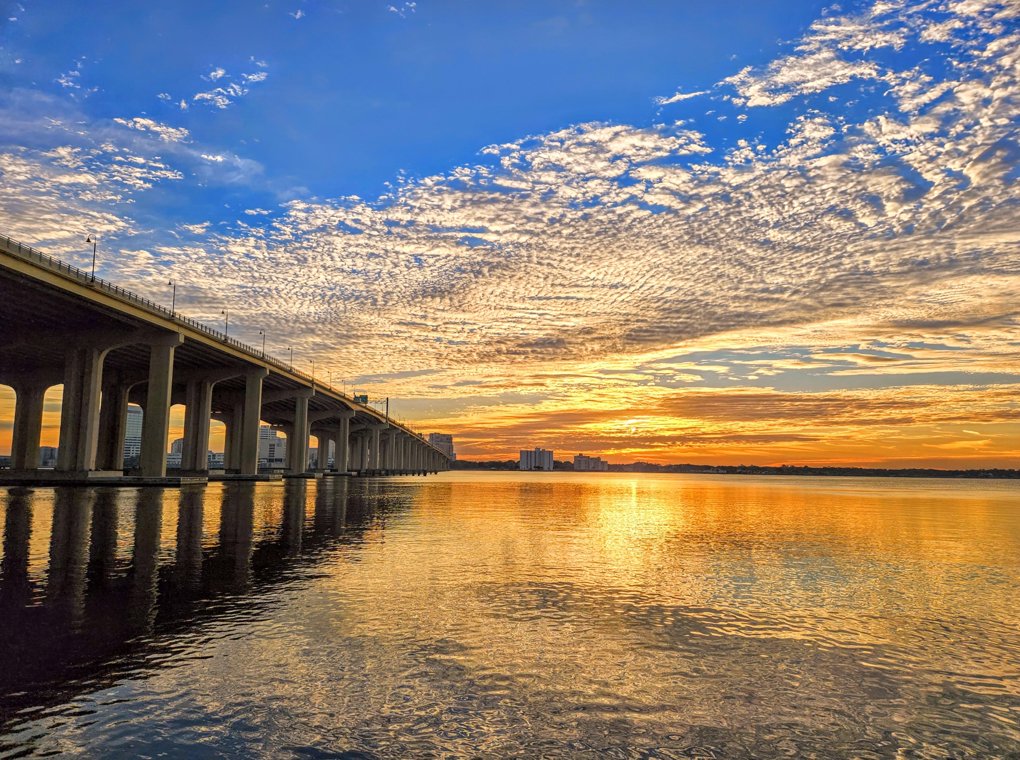 Bridge extending over calm water on the left, with a vibrant sunset casting golden reflections. Distinctive, wispy clouds spread across a blue and orange sky. Buildings appear in the distant background along the horizon.