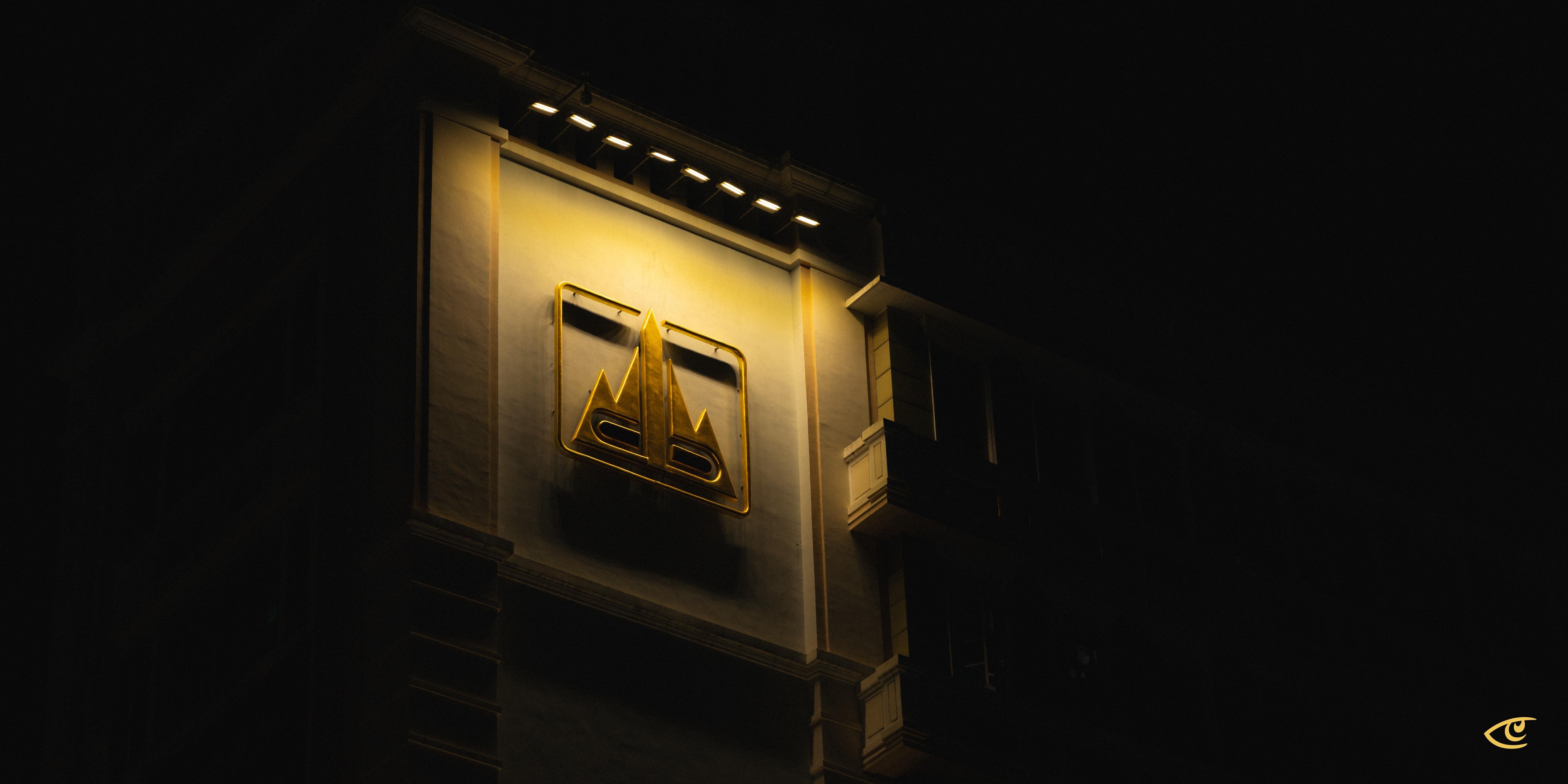 Illuminated gold emblem on a tall building façade at night, viewed from below; spotlights along the roofline cast a warm glow while the surrounding sky and edges are mostly dark.