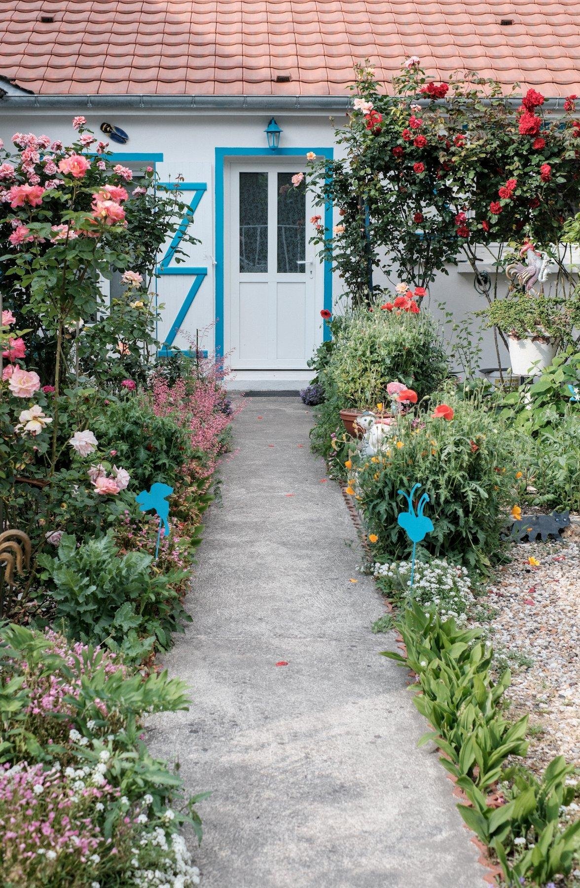 Photo verticale. Petite allée privée en béton, bordée de verdure et de fleurs, menant à une maison à la façade et porte blanche, avec le cadre de porte bleu et les renfort de volets en bois dans le même bleu. Au dessus de la porte, une lampe aussi dans le même bleu