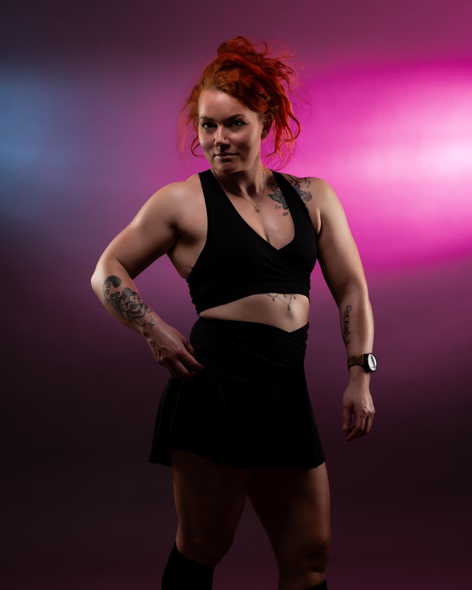The image features a person with red hair posing confidently against a backdrop with pink and blue lighting. The individual is wearing a black outfit, and you can see tattoos on their arms. The setting gives an artistic and vibrant feel.
