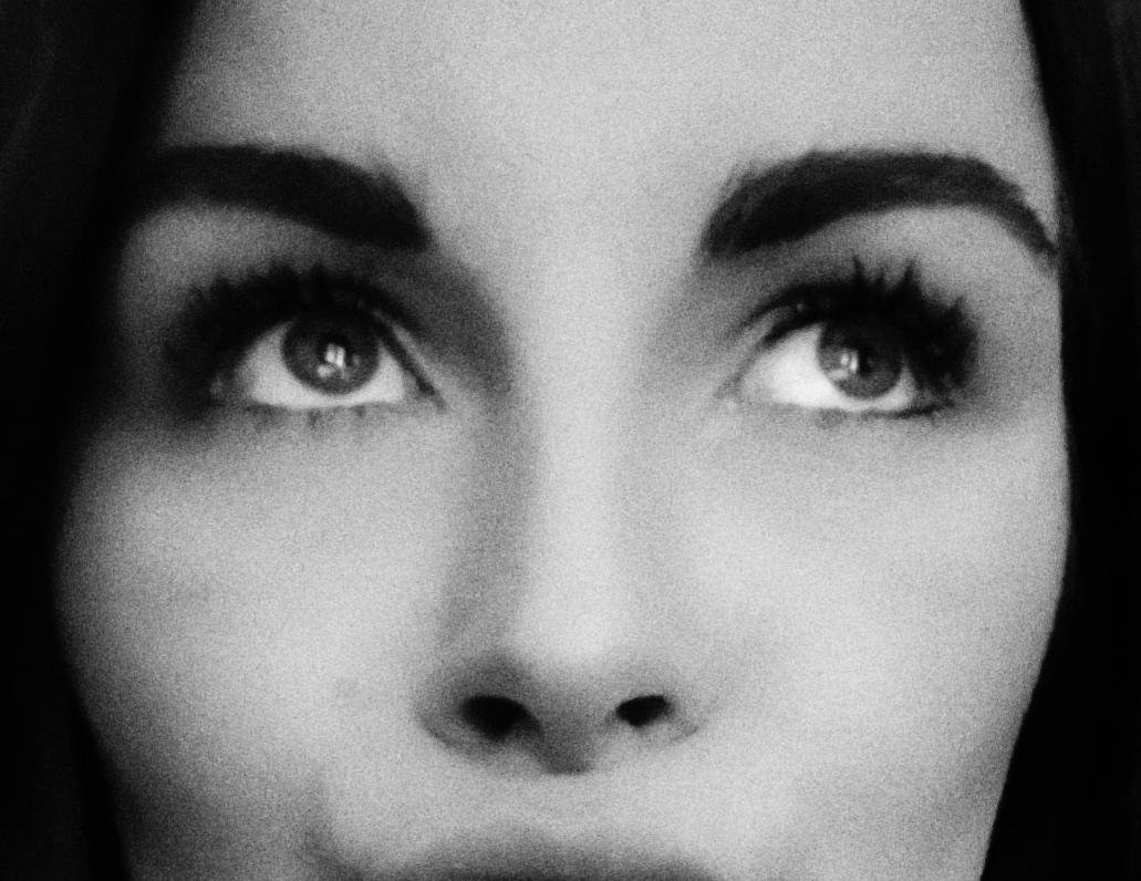 Black-and-white close-up of a woman’s face cropped at the forehead and mouth, emphasizing her eyes looking upward, thick eyebrows, and nose against a soft gray background.