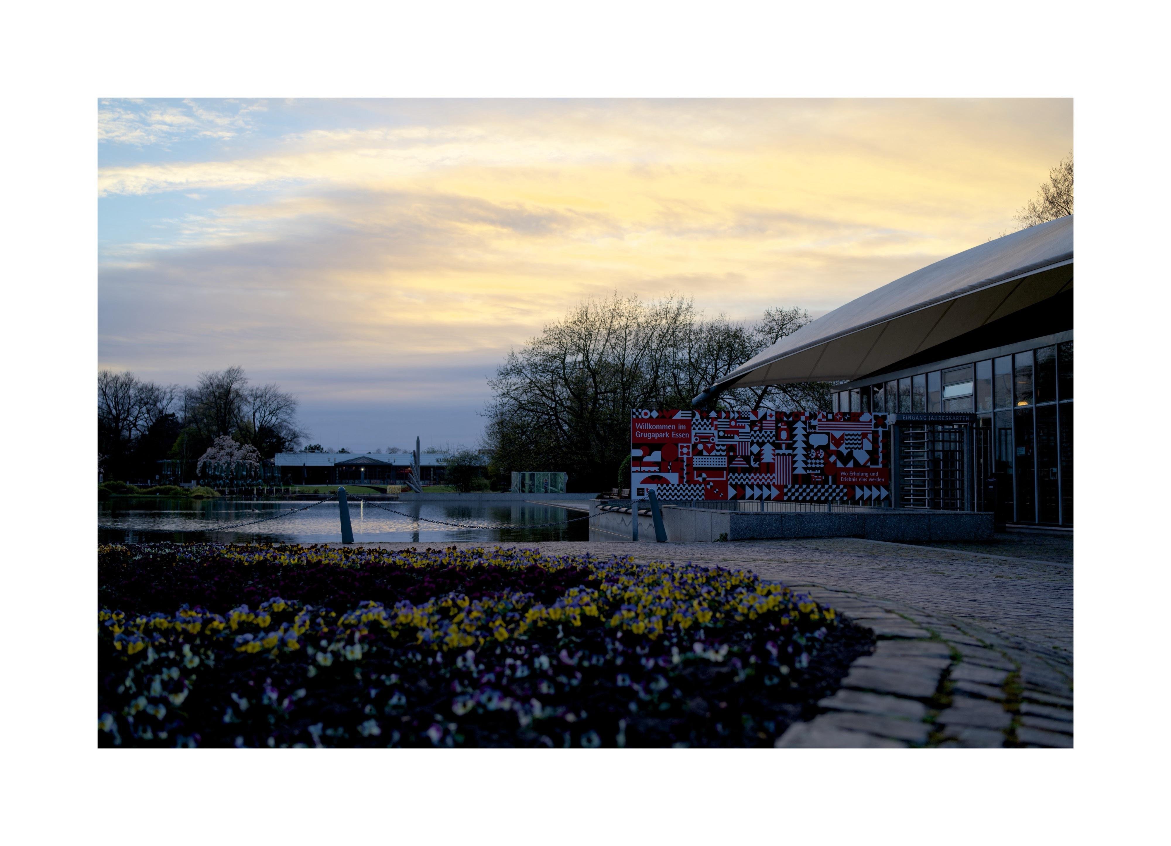 Blumenbeet im Vordergrund vor einem modernen Gebäude mit Glasfront und bunter Wandgestaltung, daneben ein ruhiger Teich unter einem gelb-blauen Abendhimmel. Auf einem roten Schild mit Blumenmuster steht "Willkommen im Grugapark Essen."