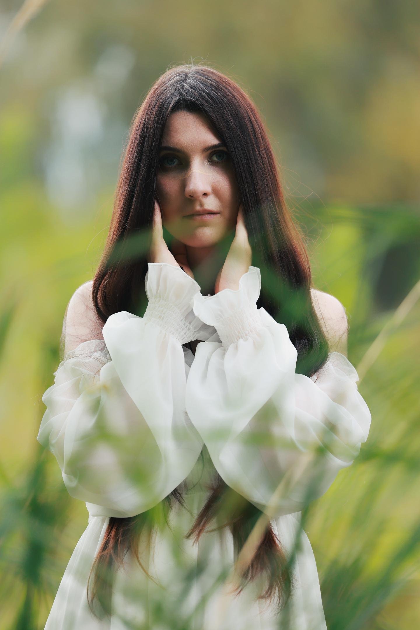Woman with long dark hair wearing a white off-shoulder dress stands amid tall grass, hands gently touching her face. Background is blurred with green and brown hues, suggesting an outdoor, natural setting.