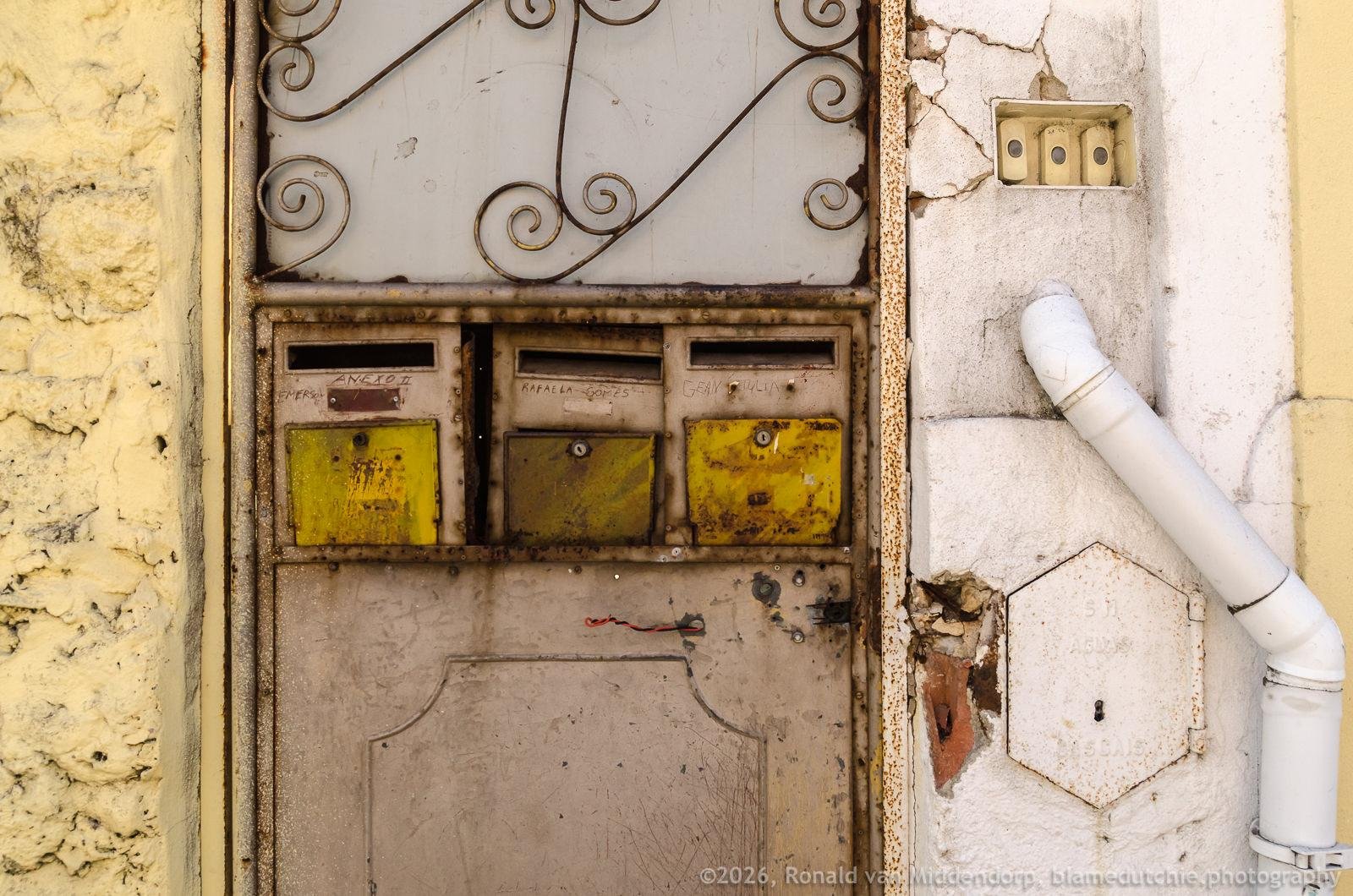 Three worn metal mailboxes with yellow labels set into a beige door with decorative wrought-iron scrollwork, beside a cracked white wall with a small electrical box and a white drainpipe.