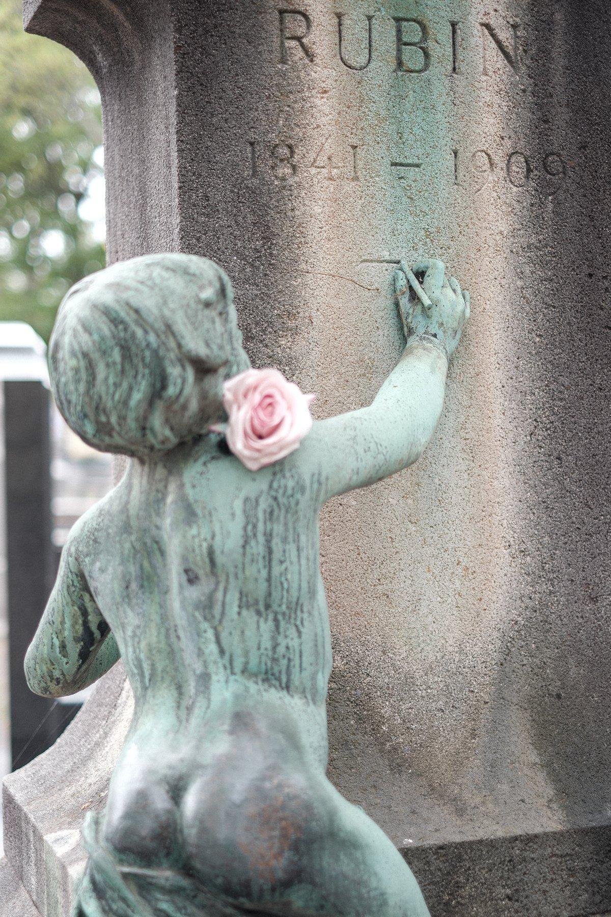 Photo verticale. Détail sur un pierre tombale. Une statue vert de gris d’un chérubin qui est en train de graver un trait sur la stèle. Une rose rose est posée sur son épaule