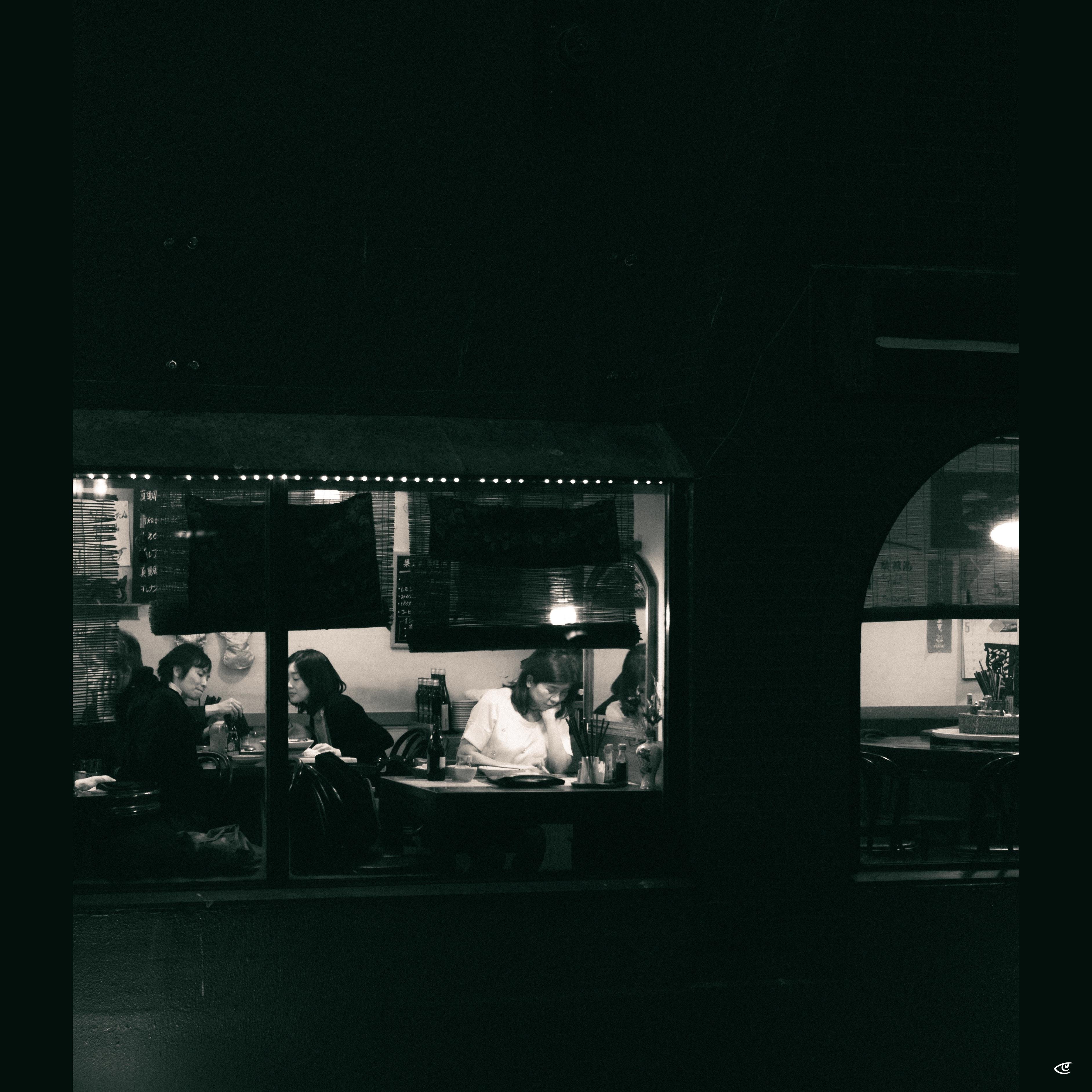Black-and-white nighttime view through a restaurant window, with several diners seated at tables inside under small string lights; dark foreground framing suggests the scene is photographed from outside.