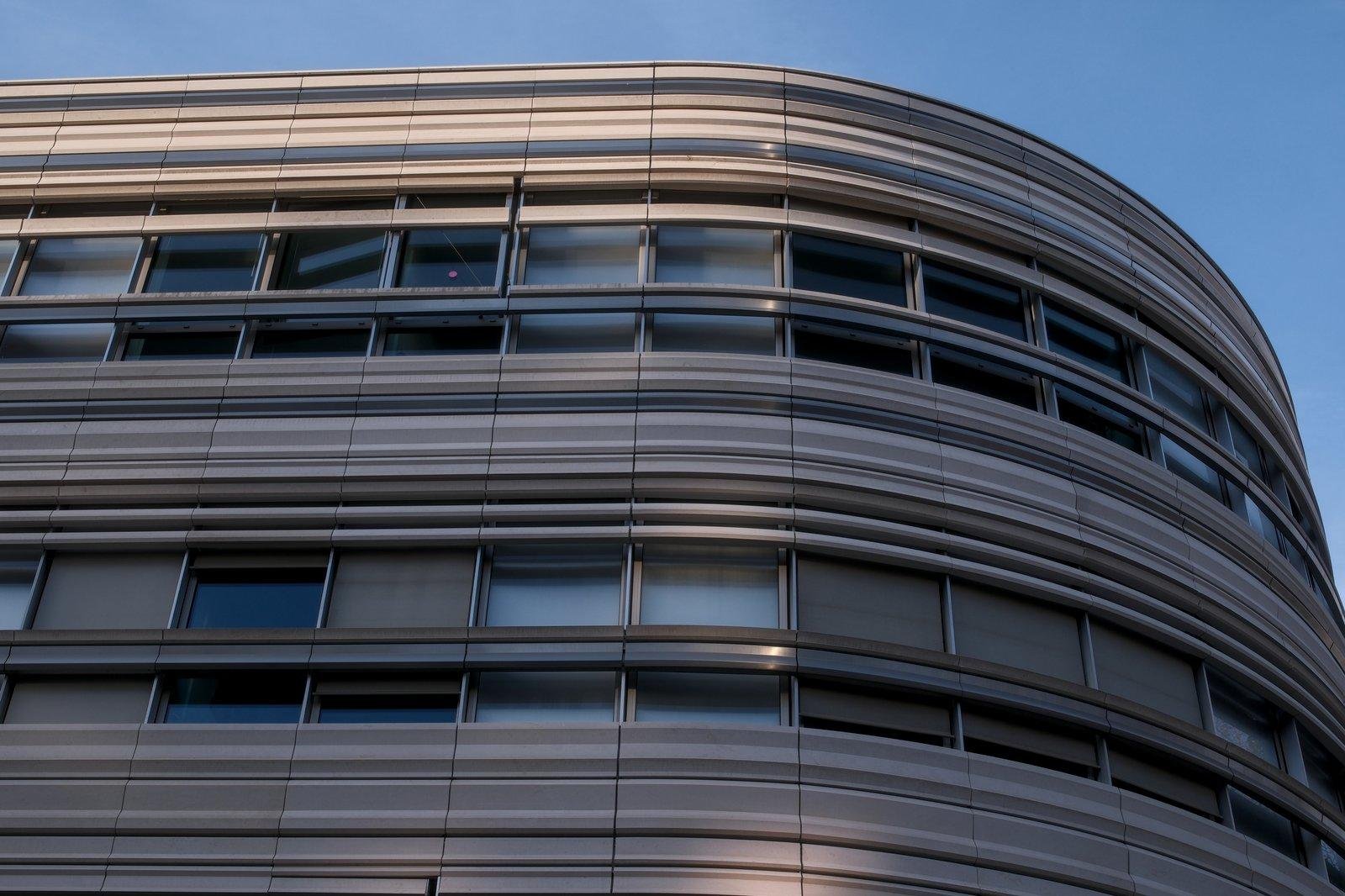 Photo horizontale. Un bout d’immeuble style années 80-90 avec plein de lignes horizontales, qui tourne sur la droite de l’image, sous un ciel bien bleu