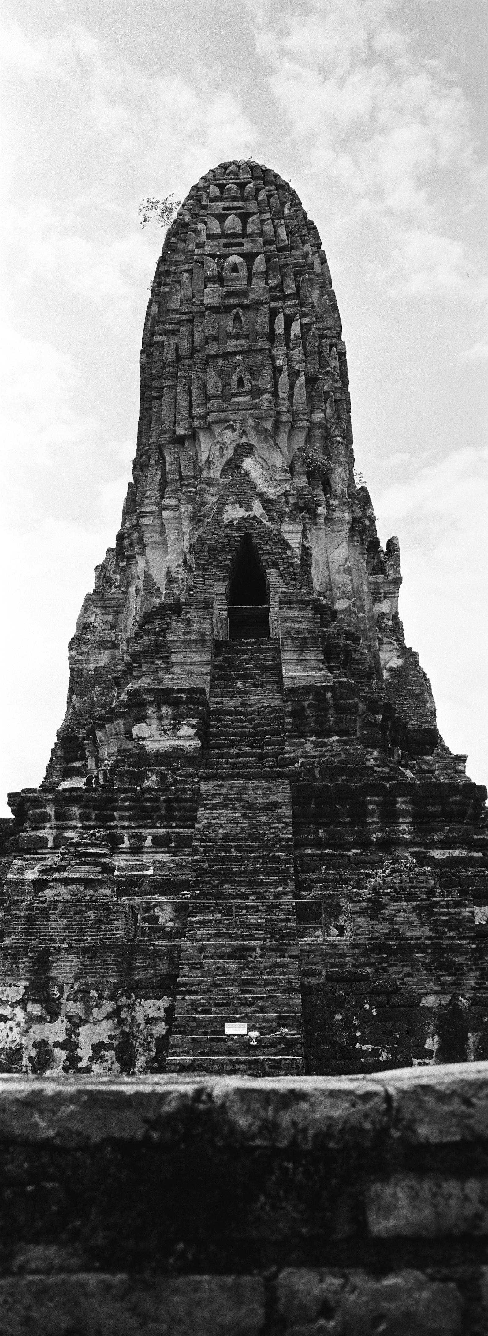 Black-and-white panoramic vertical photo of a tall, weathered stone temple tower with a central staircase leading to a dark arched doorway, set against a cloudy sky