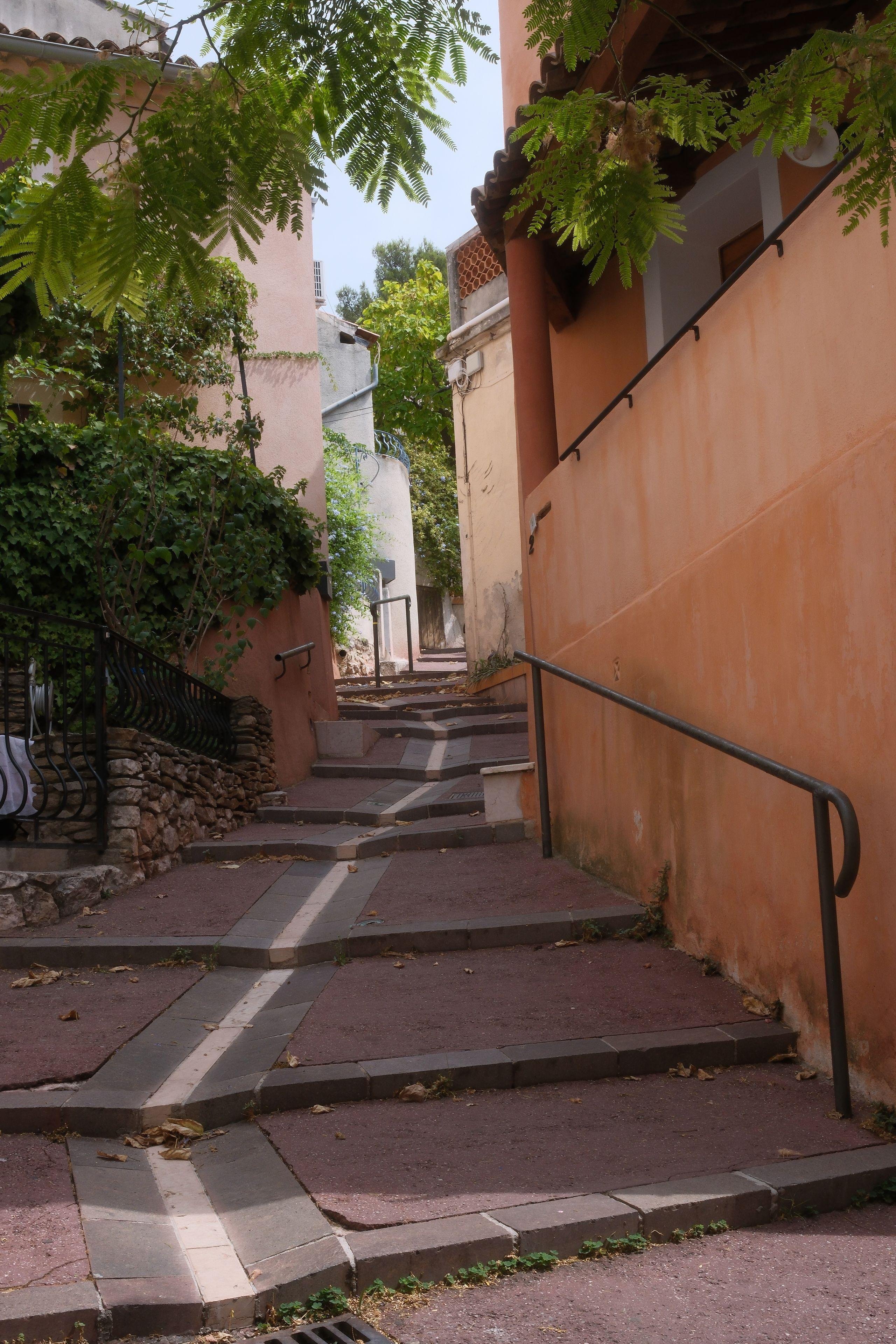 Photo verticale. Une allée piétonnes faite de marches profondes et larges, qui ont toutes une rigole pour l’écoulement de l’eau. Une rampe est sur la droite devant un mur orange.