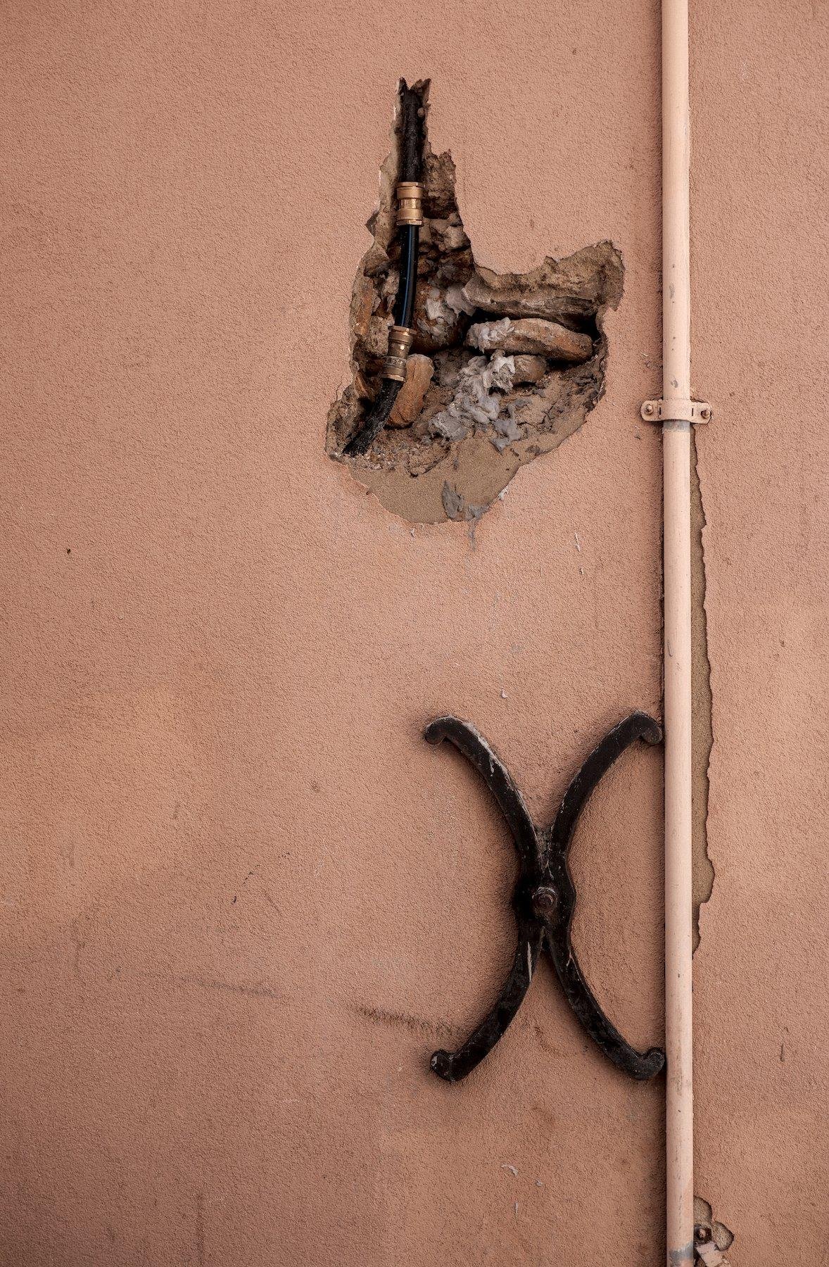 Photo verticale. Un mur saumon barré verticalement d’une gaine électrique peinte, une décoration en fer forgé en forme de X arrondi, et en haut, un trou dans le mur laissant apparaître un tuyau noir avec un liseret bleu, probablement de gaz, avec 2 jointures dorées