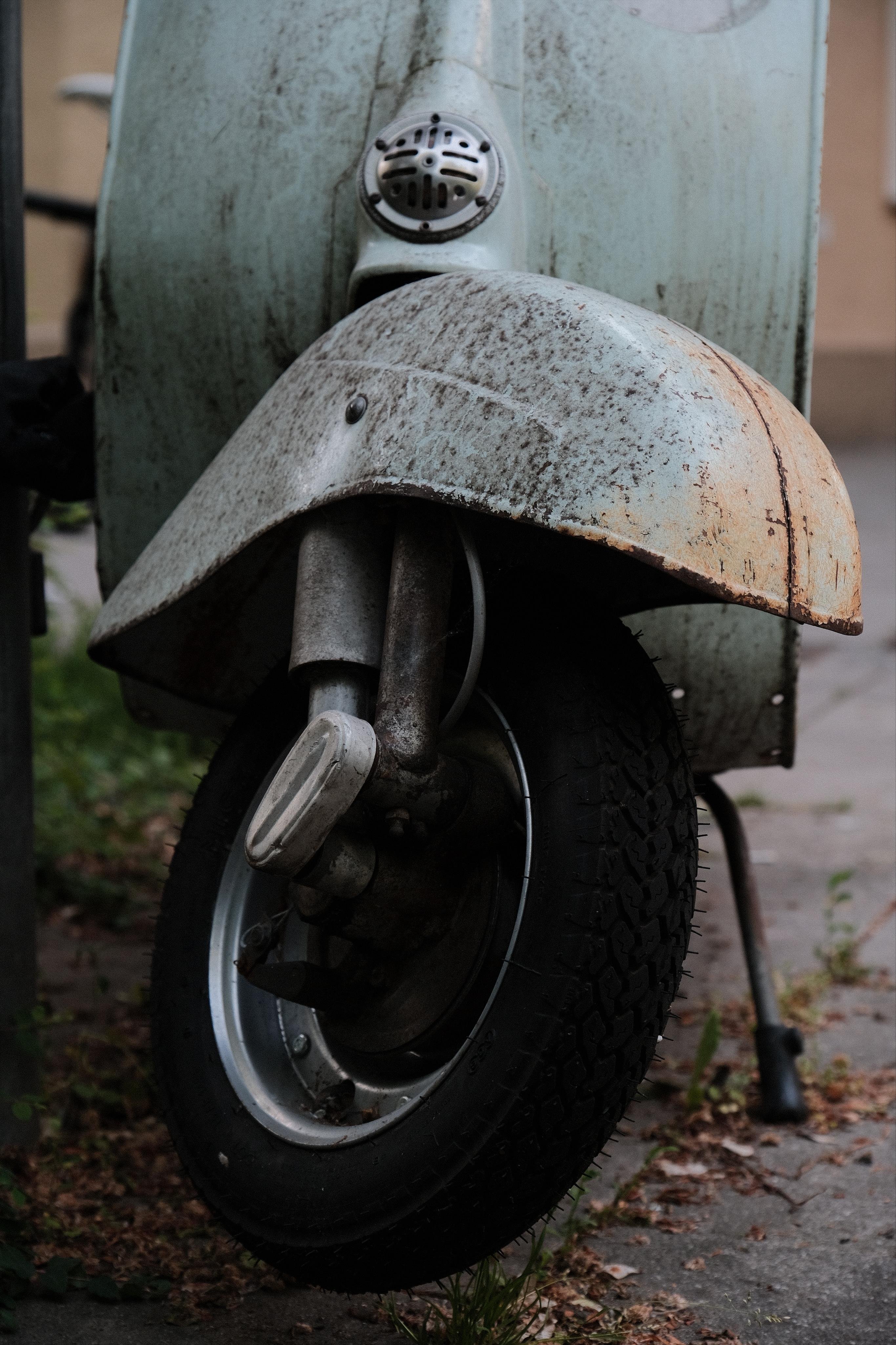Close-up of the front wheel and fender of an old scooter, showing a worn, rust-speckled light-blue fender above the wheel that is at a 45 degree angle to the camera. The scooter stands on a paved surface with scattered grass.