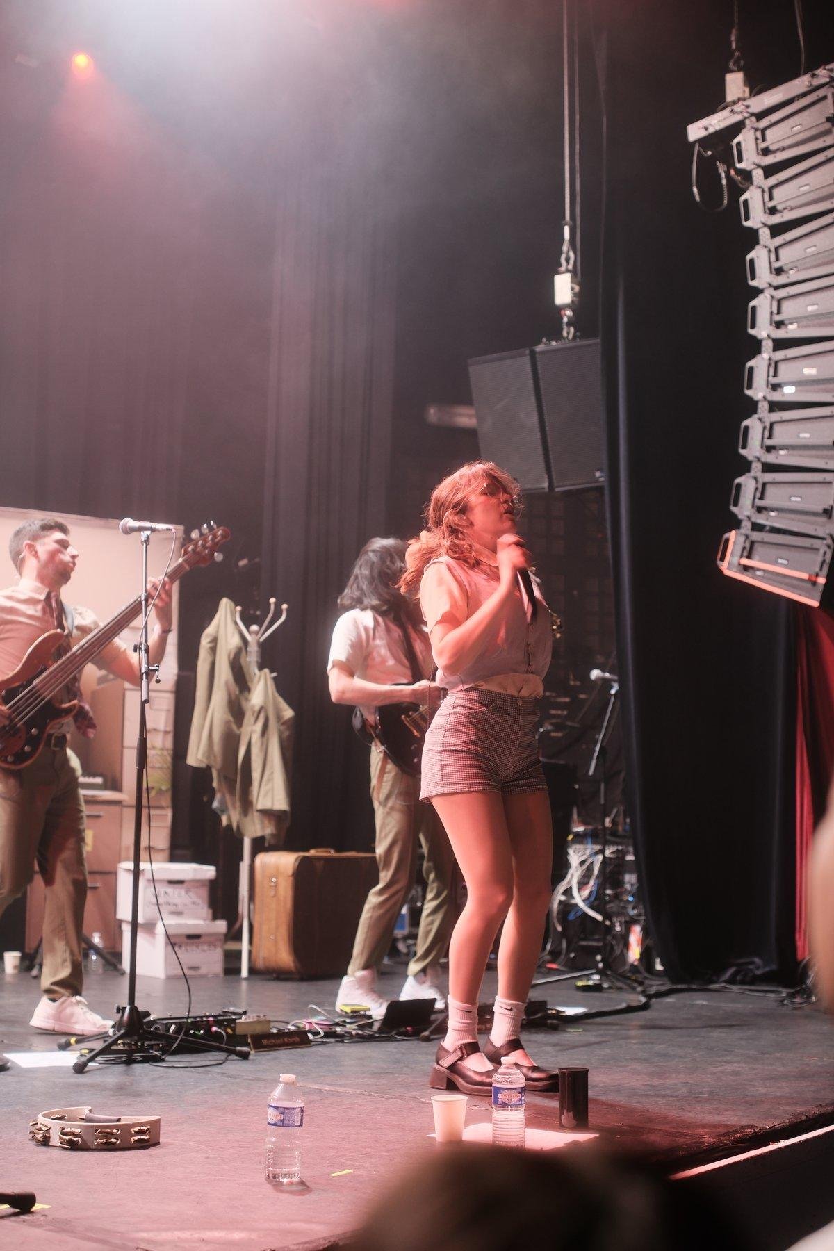 Photo portrait de Gracie Lawrence du groupe Lawrence, les yeux fermés, en train de danser, en transe. Derrière elle on aperçoit le bassiste et le guitariste concentrés. La lumière éclaire la fumée de la scène depuis le coin haut gauche.