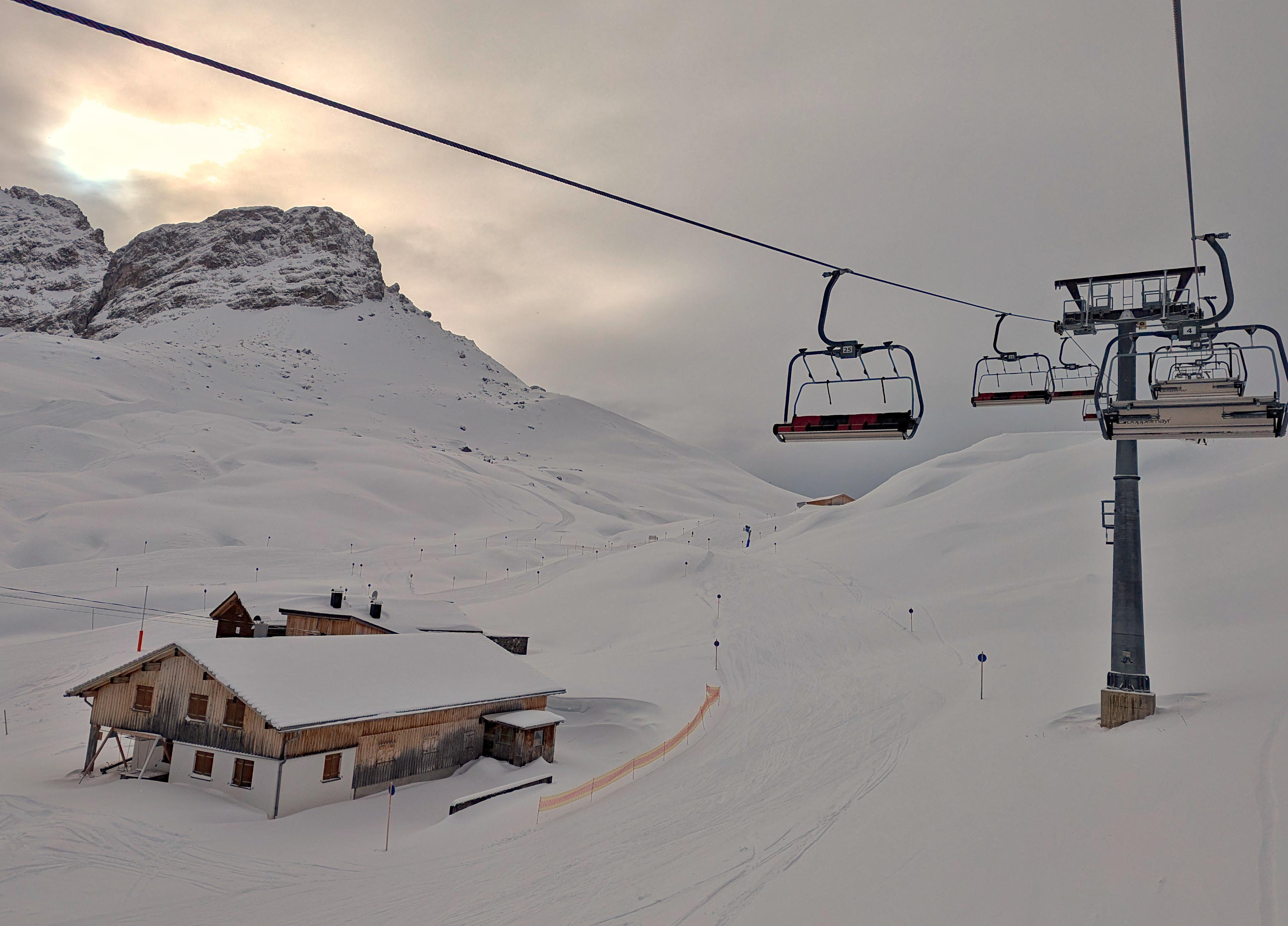 Snow-covered ski resort with a chairlift line crossing the top of the frame, two empty chairs midair, and a lift tower on the right; a wooden lodge sits partly buried in snow in the lower left, with groomed ski tracks leading toward distant snowy mountains under a cloudy sky.