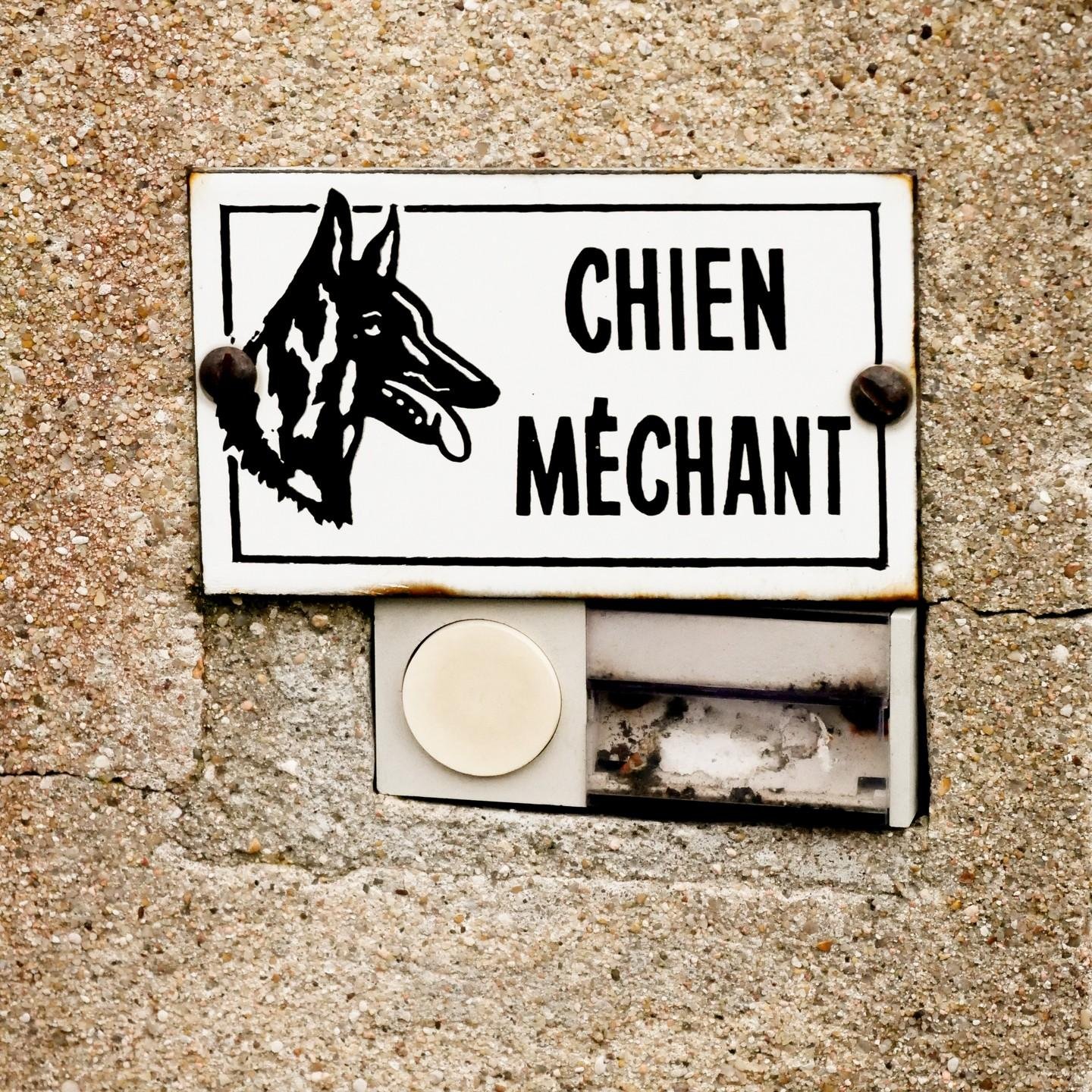 This image shows a sign with the text "CHIEN MÉCHANT," which is French for "Beware of Dog" or "Bad Dog." There is an illustration of a dog's head on the sign, and below it is a doorbell button mounted on a wall.