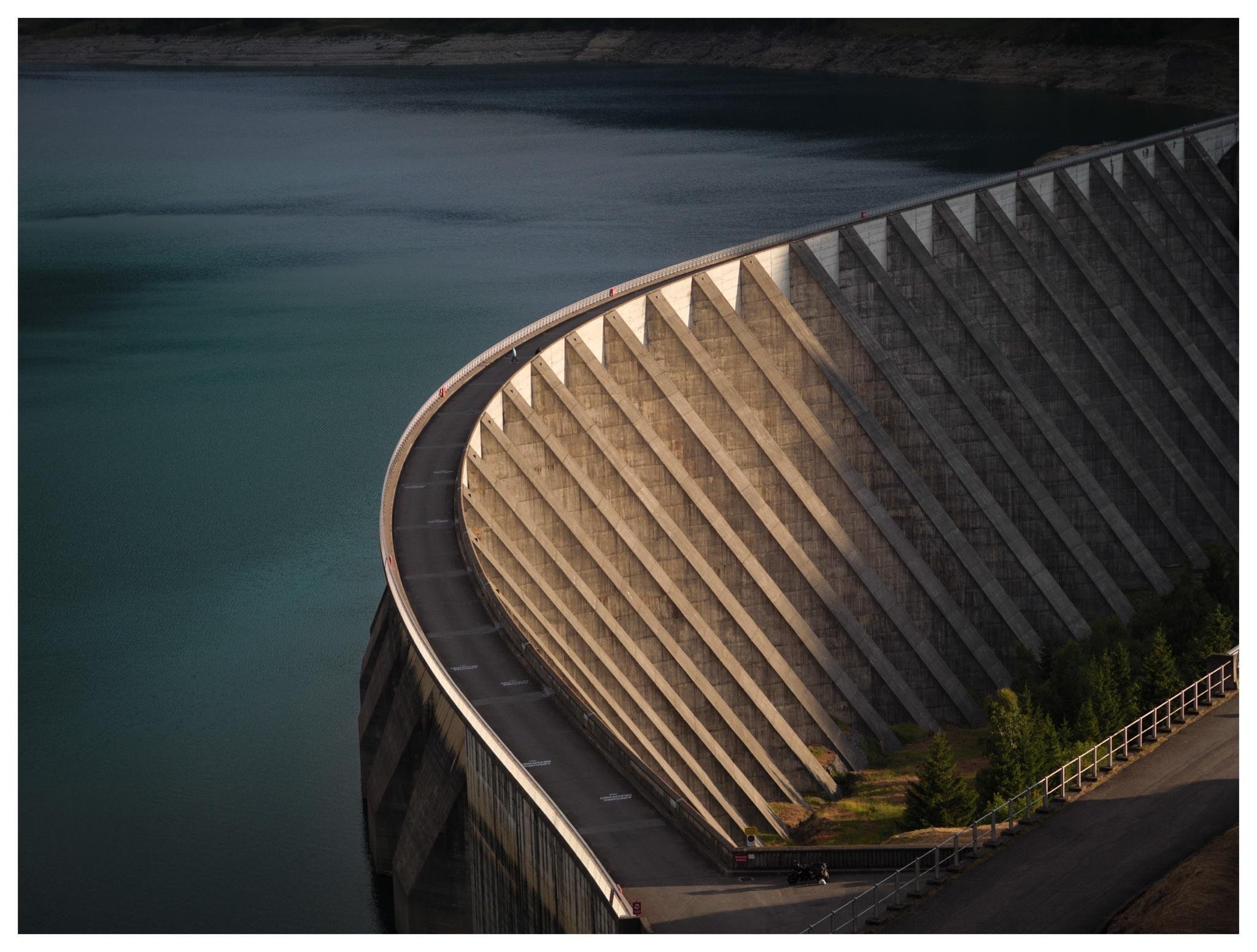 Curved concrete dam wall with repeating vertical buttresses, a narrow roadway along the top, and turquoise reservoir water on the left; view from above with a small access area and railing at the lower right.