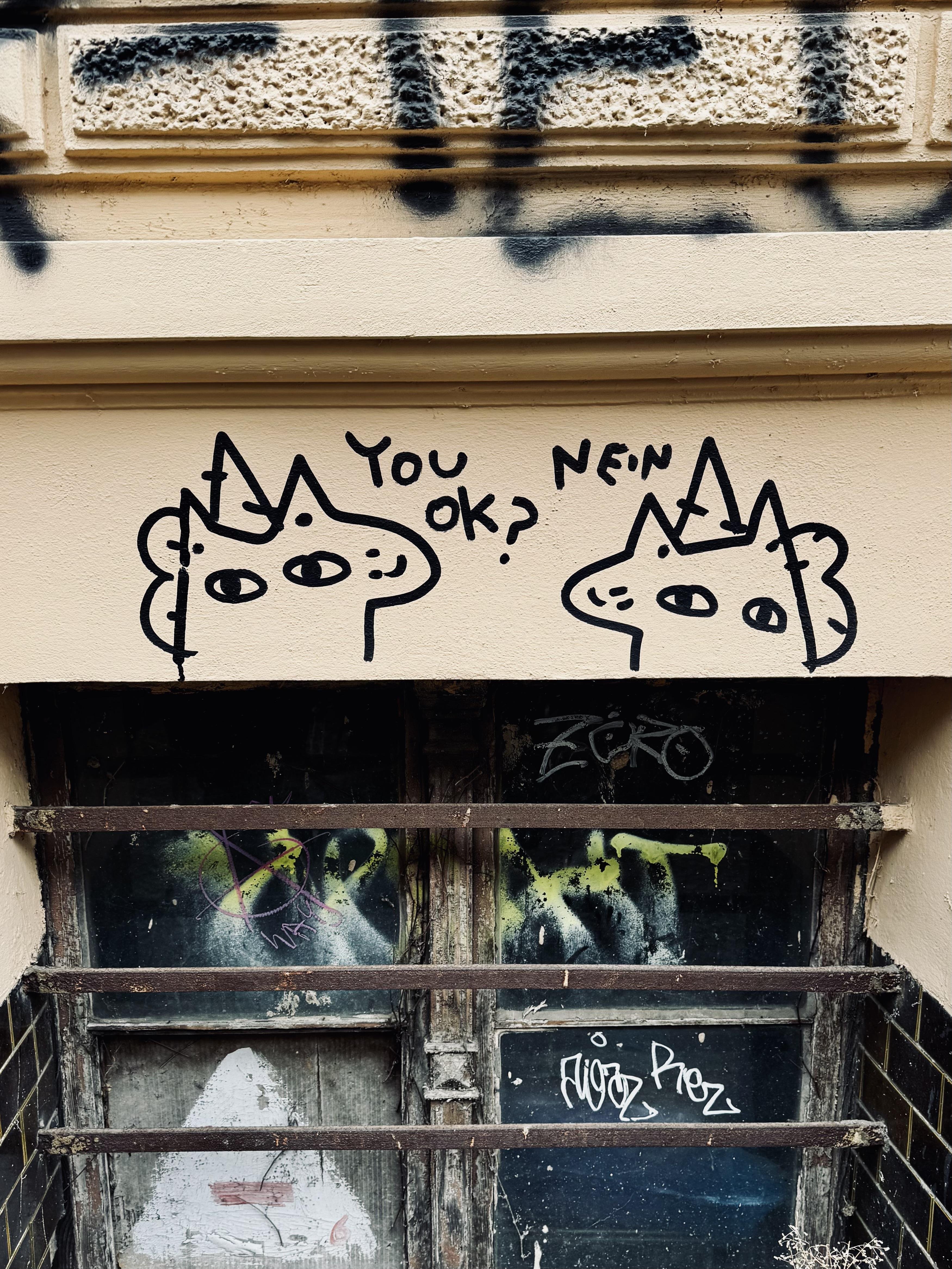 The image depicts a piece of street art featuring two cartoon-like faces with horns resembling unicorns. One of the figures is asking "YOU OK?" and the other responds with "NEIN," which means "no" in German. The artwork is drawn on a wall, and there is graffiti visible underneath on a boarded-up window.