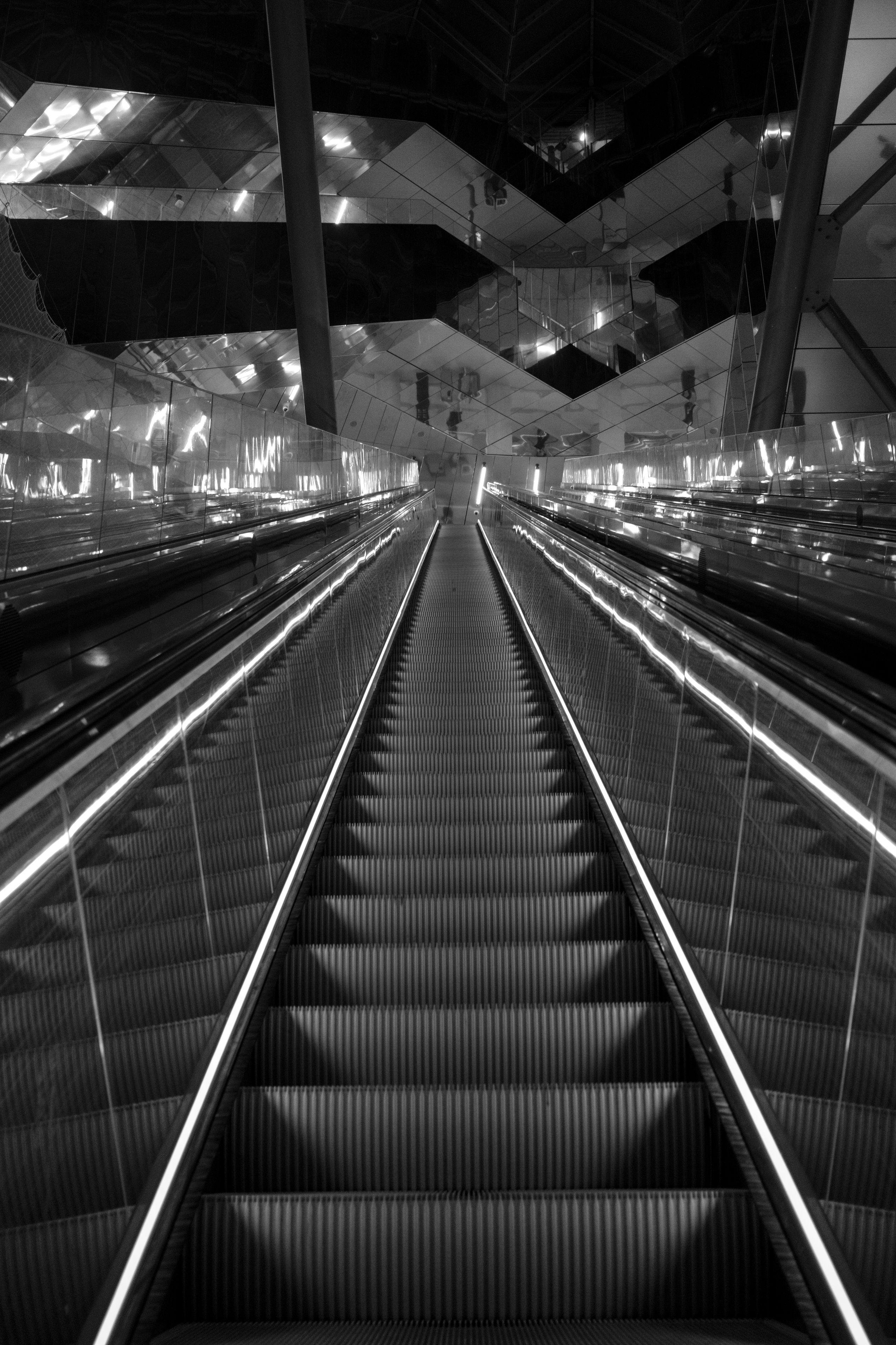 Photo verticale en noir et blanc. Un très long escalator qui monte, le plafond est fait de surfaces réfléchissantes faisant perdre ses repères (peut être qu’on descend en fait ?)