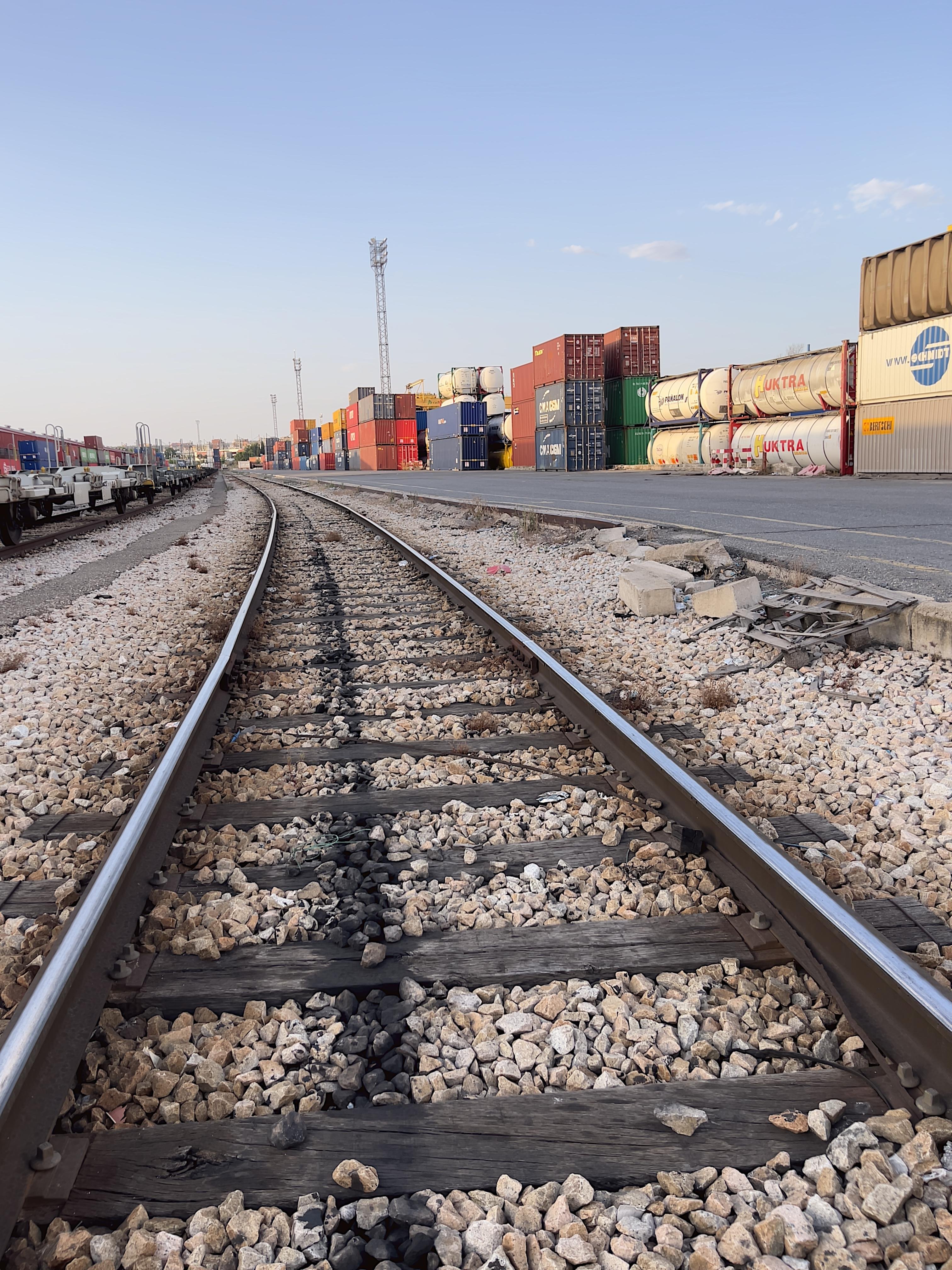 Railroad tracks curve to the left, bordered by gravel and wooden ties. Stacked shipping containers in various colors line the right side, with some railcars visible on the left. A clear blue sky is overhead with a few clouds and tall industrial lights are positioned in the distance.