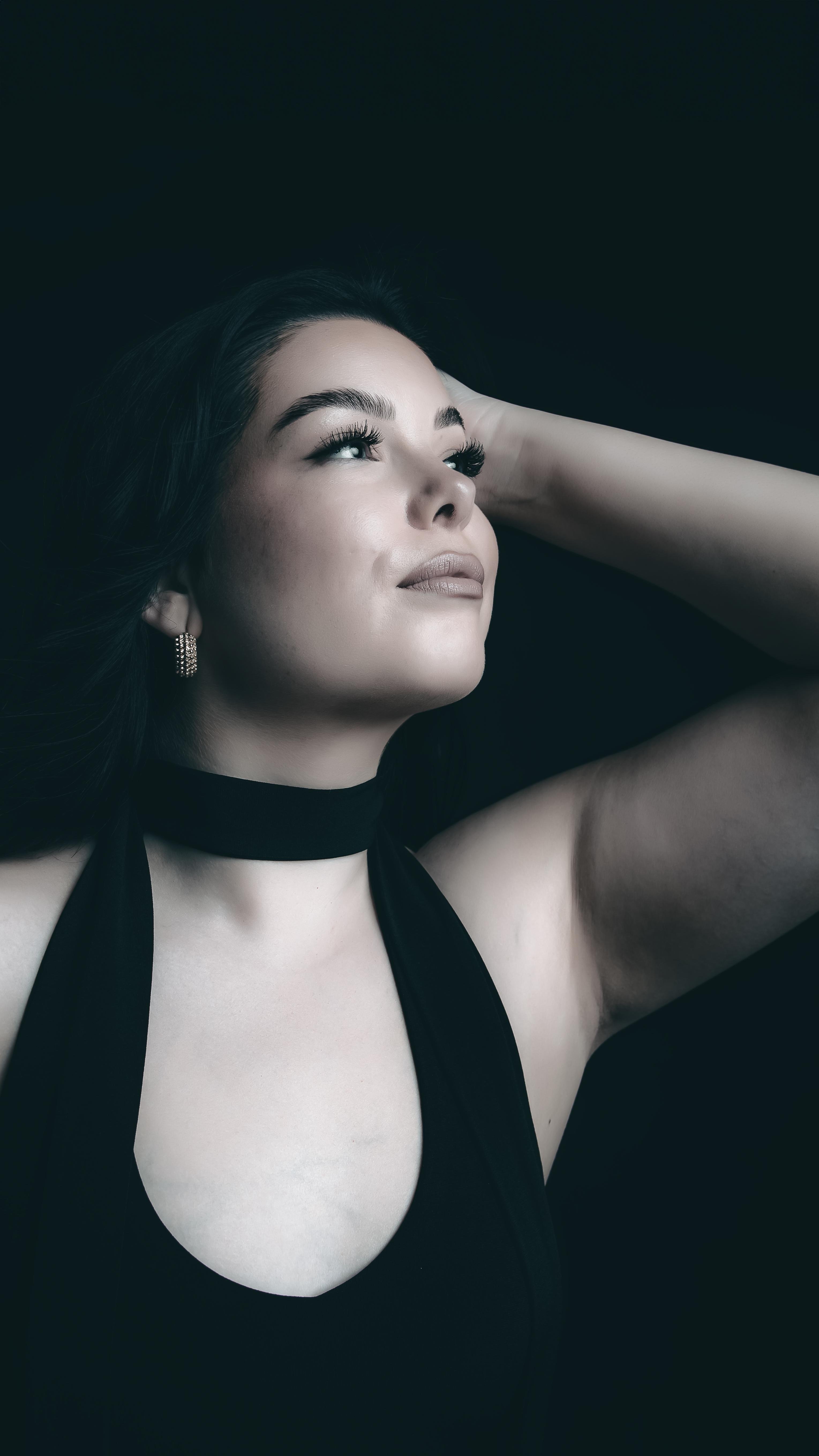 The image shows a woman with dark hair, wearing a black top. She has her arm raised, touching her head. The background is dark, highlighting her face and features.