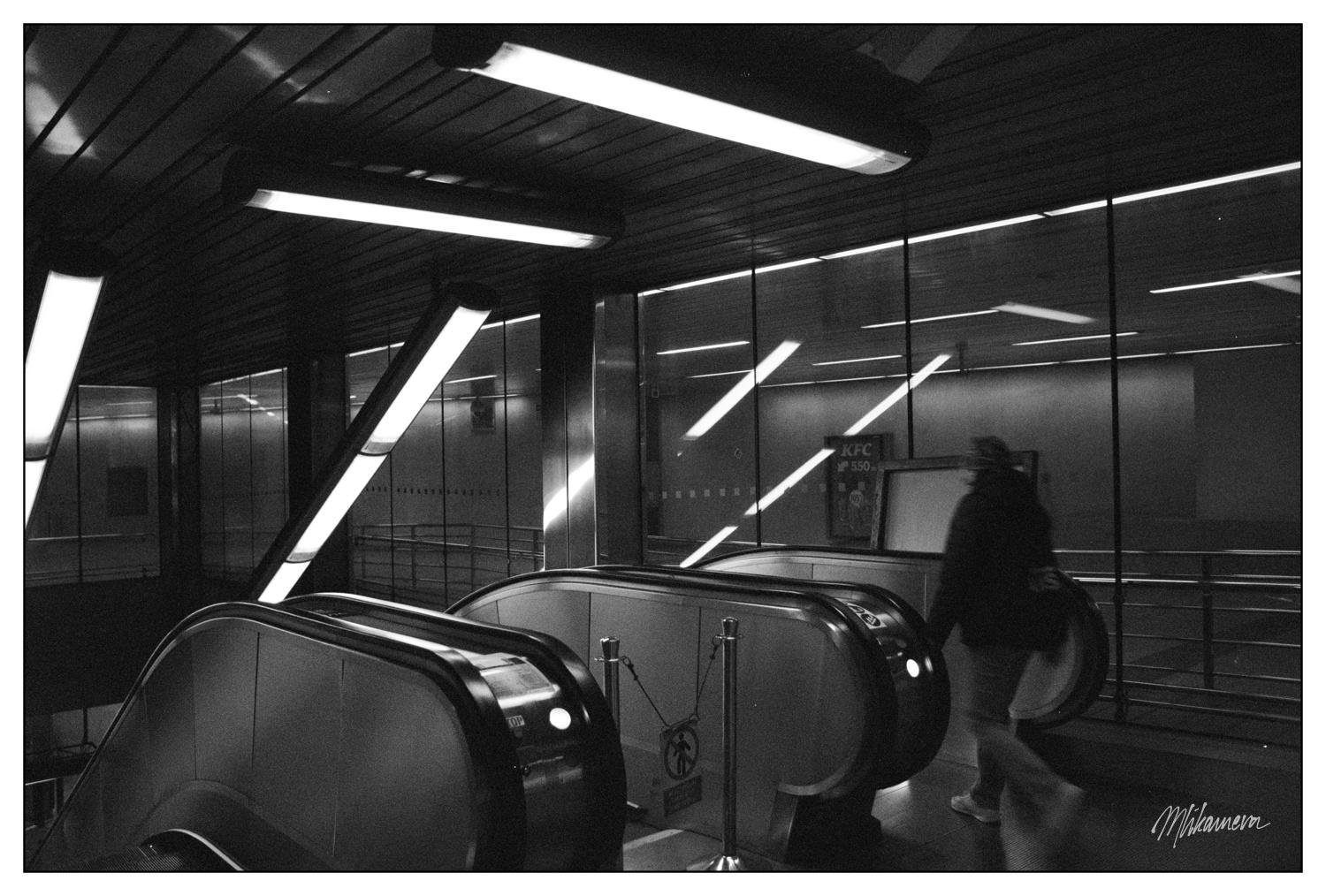 A monochrome photo of a underground entrance to escalators with fluorescent lamps hanging from the ceiling and a person walking towards the escalators