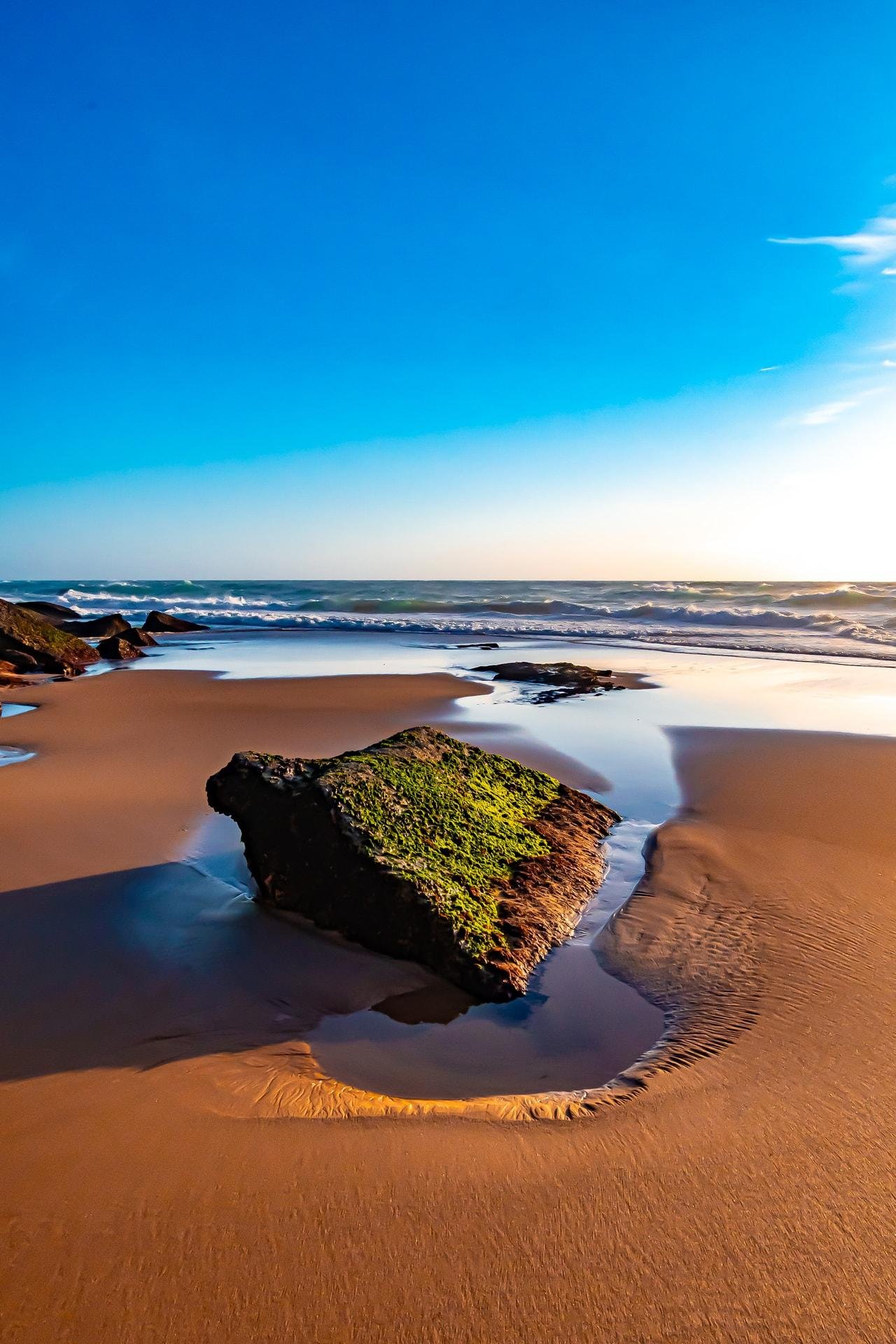 Sandy beach with a large, moss-covered rock in the foreground surrounded by shallow water. Waves can be seen in the distance under a clear blue sky with sunlight illuminating the horizon.