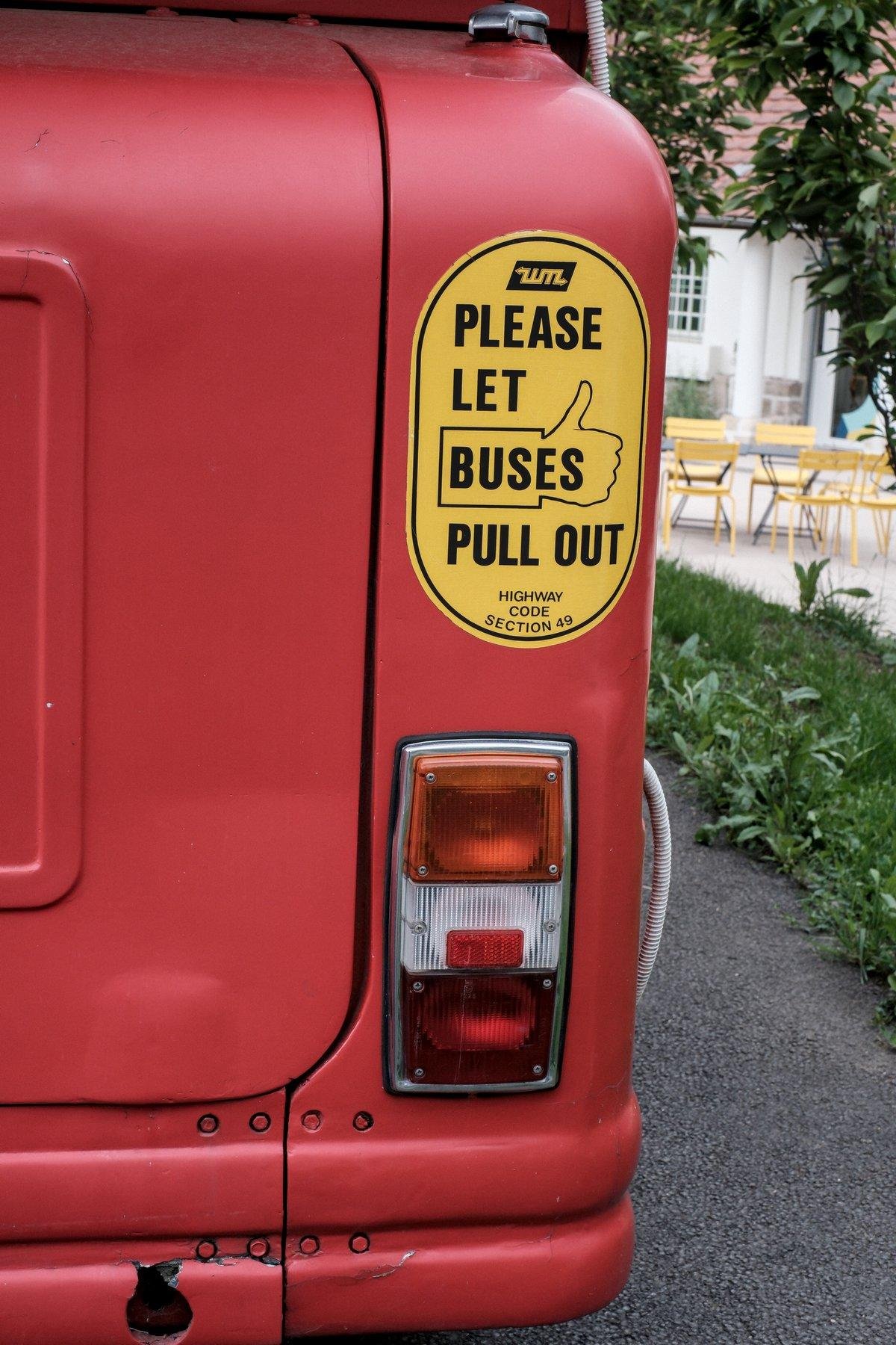 Photo verticale. Vu du coin arrière droit d’un bus londonien rouge, avec le feu arrière et un sticker jaune sur lequel est écrit en noir « Please les buses pull out »