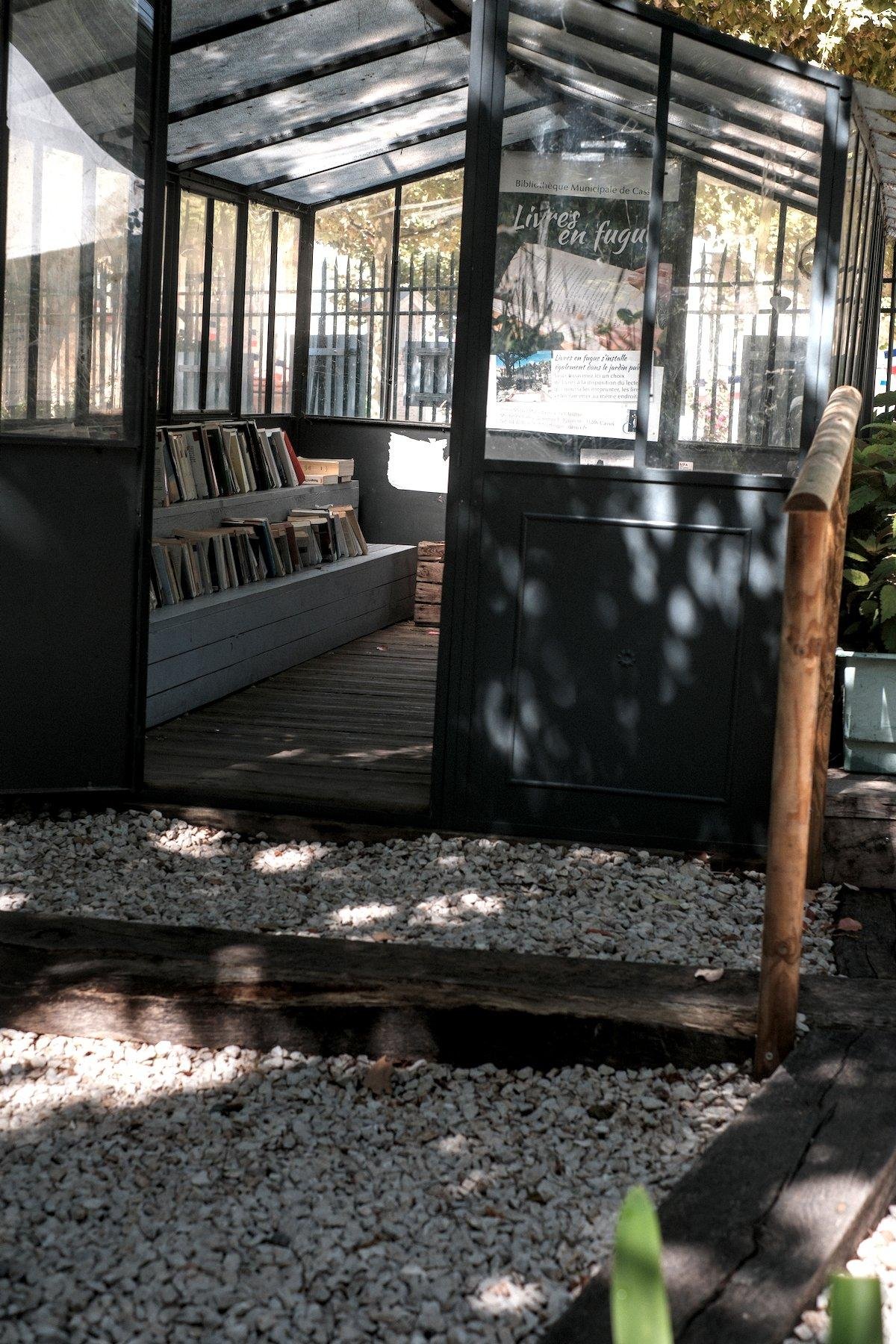 Photo verticale. Une petite véranda/serre dans laquelle se trouve une petite bibliothèque publique d’échange de livres