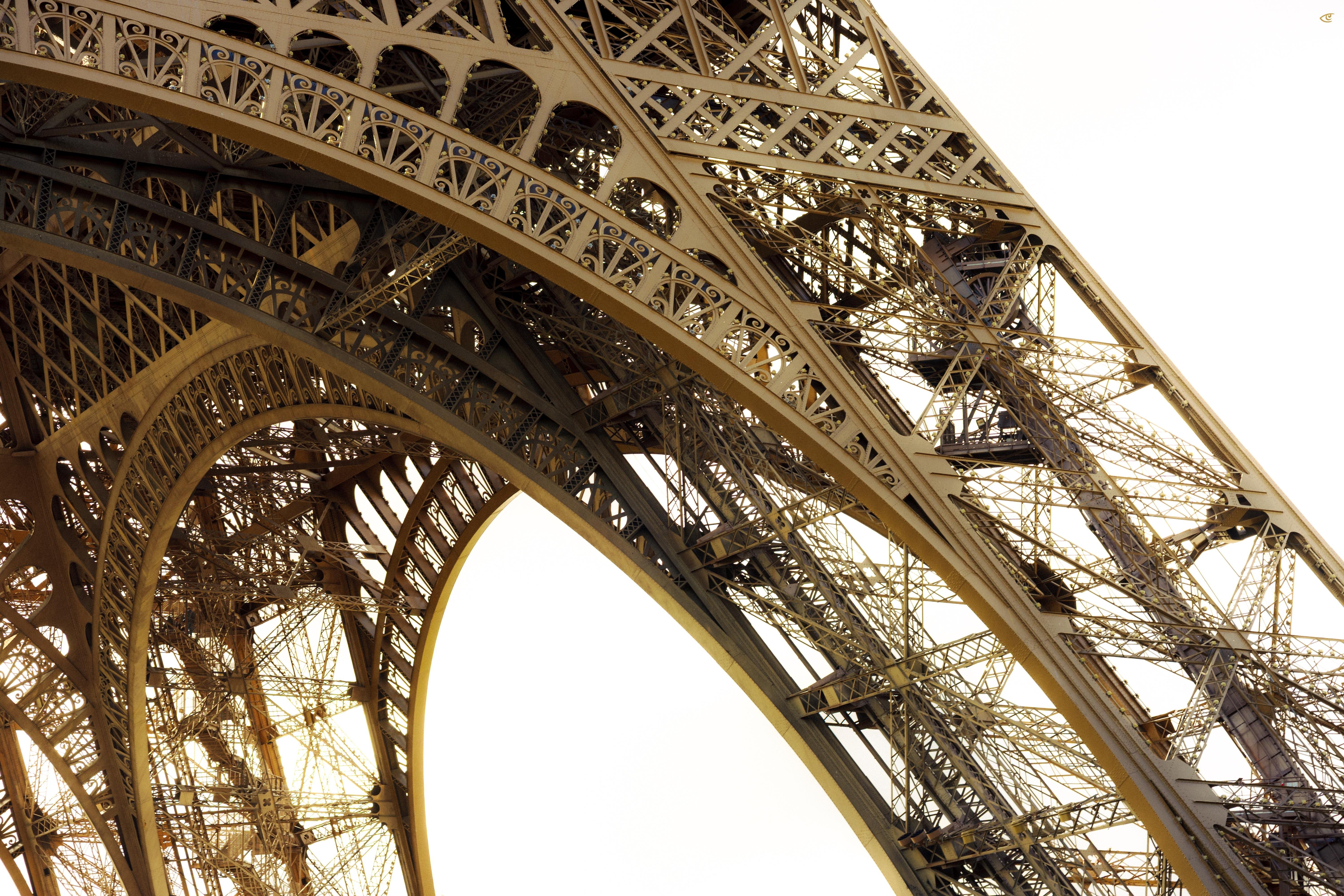 Close-up view of the Eiffel Tower’s iron lattice structure, showing intersecting beams and curved arches against a bright sky.