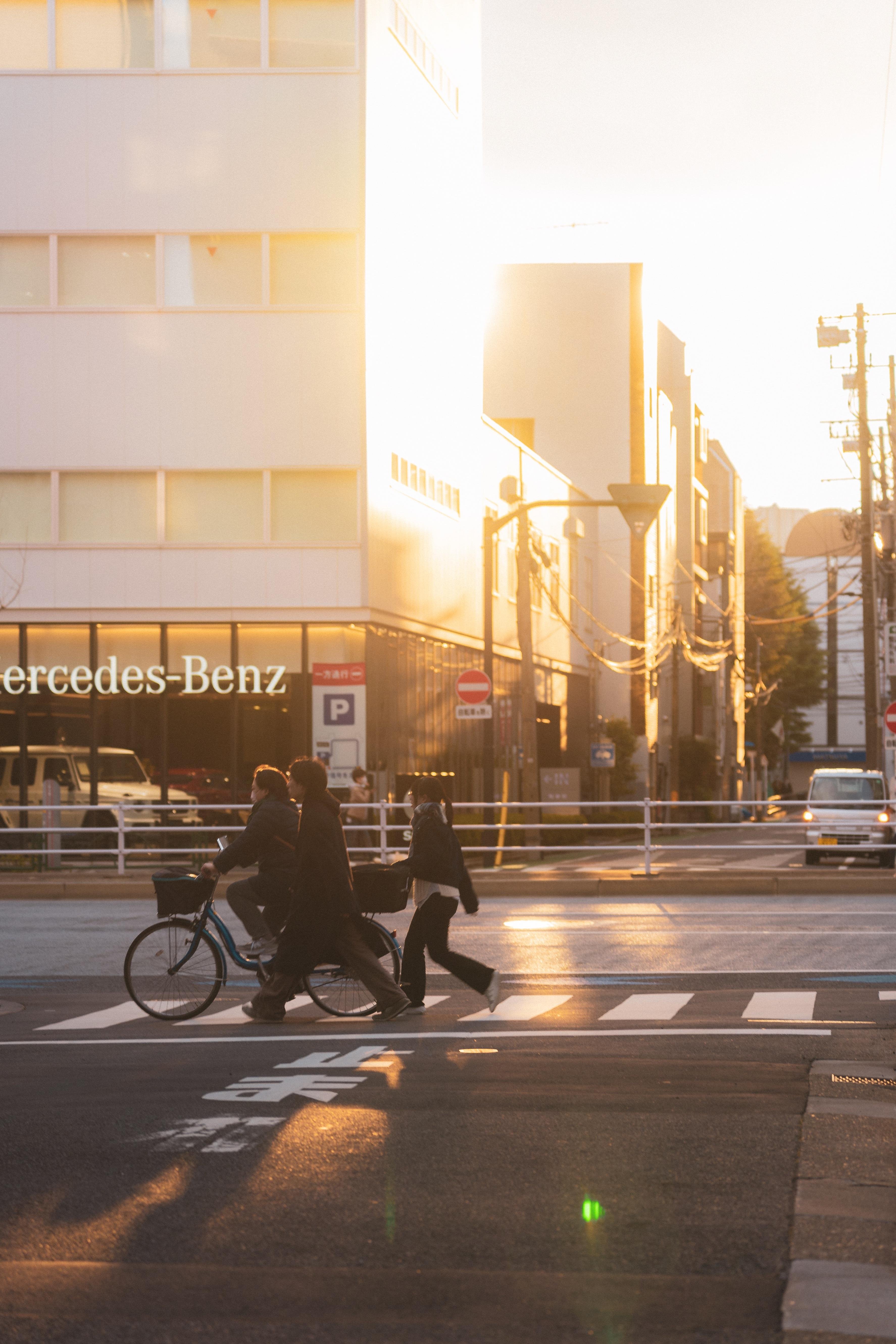 The image shows a street scene during sunset. There are people crossing a crosswalk, one riding a bicycle and others walking. In the background, there is a building with the "Mercedes-Benz" logo on it. The sunlight casts long shadows and creates a warm glow on the scene.
