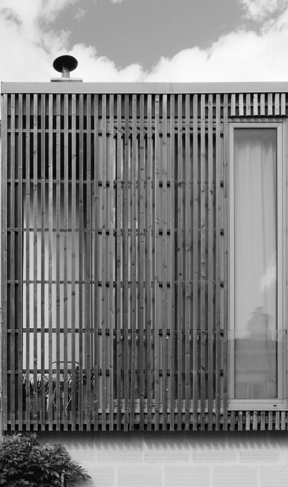 Photo format portrait en noir et blanc, montrant un bout de façade de maison, avec une baie vitrée, couverte d’une grille en bois, découpée autour de la fenêtre la plus à droite de la baie