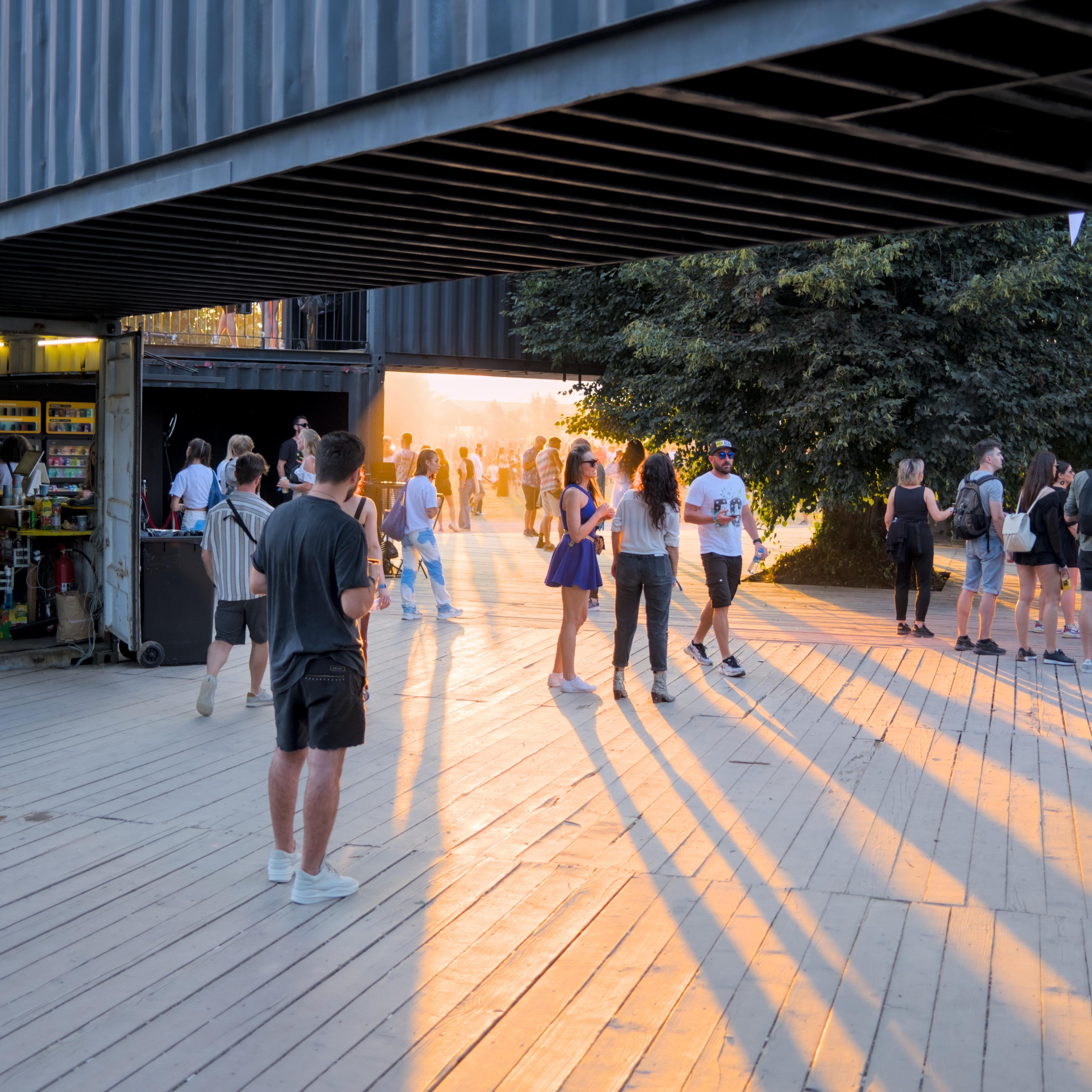 The image depicts a group of people gathered in an outdoor area with wooden flooring. There is a structure above made of shipping containers, and people are interacting and walking around. The sunlight is casting long shadows on the ground, creating a warm atmosphere. Some people are near a small vendor or kiosk.