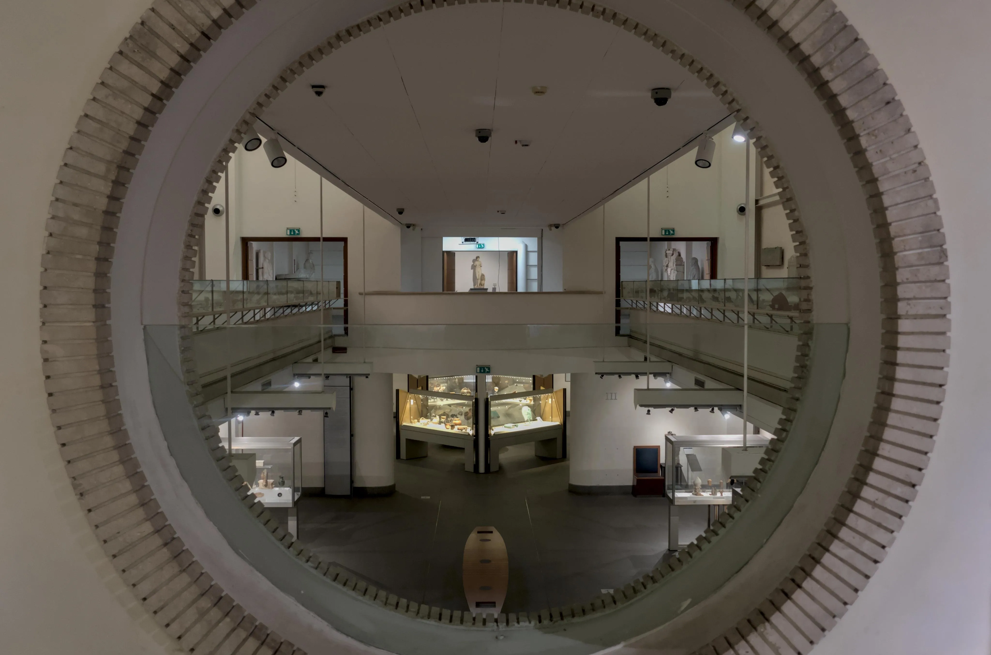 Photograph of a museum room seen through a round window. The window opens to a room with 3 floors. The window with the point of view opens to the middle floor. The bottom floor has glass case displays with smaller artifacts such as ceramics. The middle floor ends with doors to a room with life-size statues, including a statue of an armless Venus in the center of the perspective and the center of the photograph. The top floor can only be seen from underneath, as a walkway bridging the room above the window and the room above the statue of Venus. The ceiling of the multi-floor room is unseen. Overall, the architecture and display of the museum are marked by an important symmetry within the perspective of the photograph.