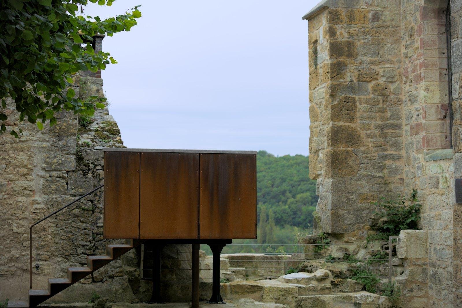 Photo paysage d’un mur ancien en ruine, avec devant le trou, une plateforme d’observation en métal rouillé, avec des escalier en métal aussi menant à la plateforme depuis la gauche de celle-ci. Des feuilles d’arbre vertes entrent dans le cadre depuis le coin haut gauche