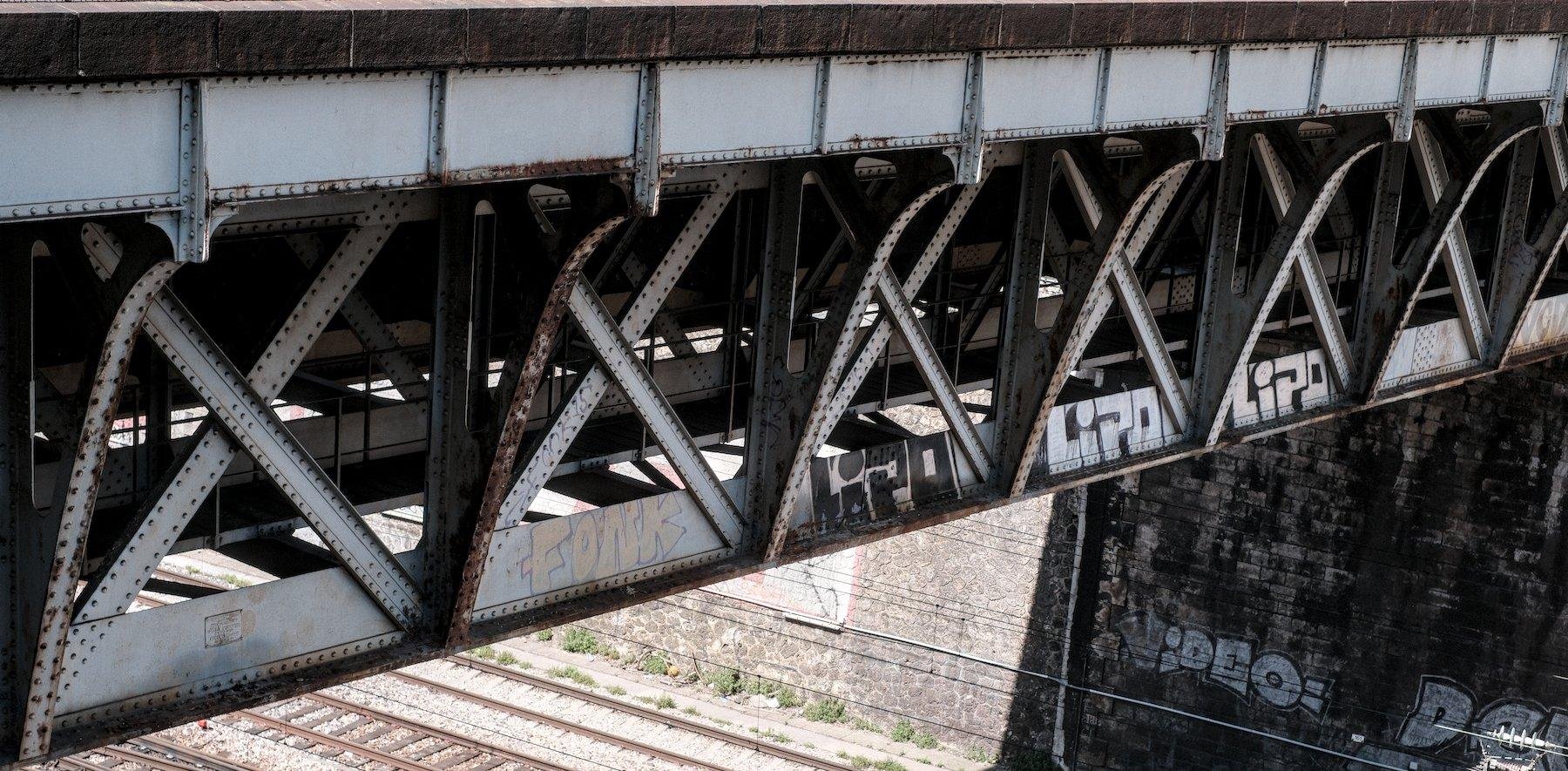 Photo horizontale. Structure en acier gris du dessous d’un pont passant au dessus de voies ferrées. Au centre, des tags