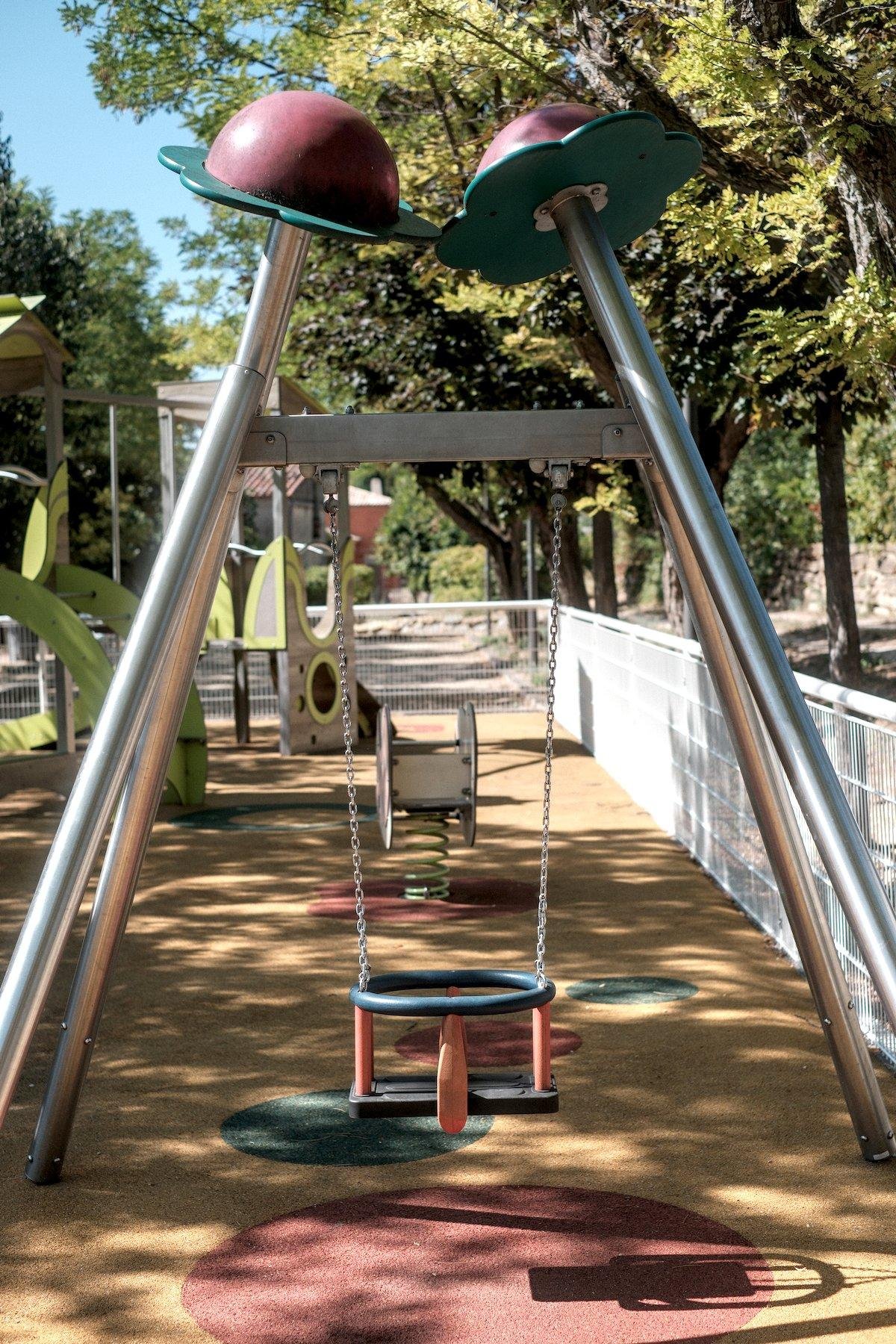 Photo verticale. Vue sur une balançoire pour petits enfants vu que le siège est une nacelle sécurisée, pendu par 2 chaînes sur une structure métallique avec les sommets en forme d’une sorte de fleur aux pétales vertes et le centre rouge
