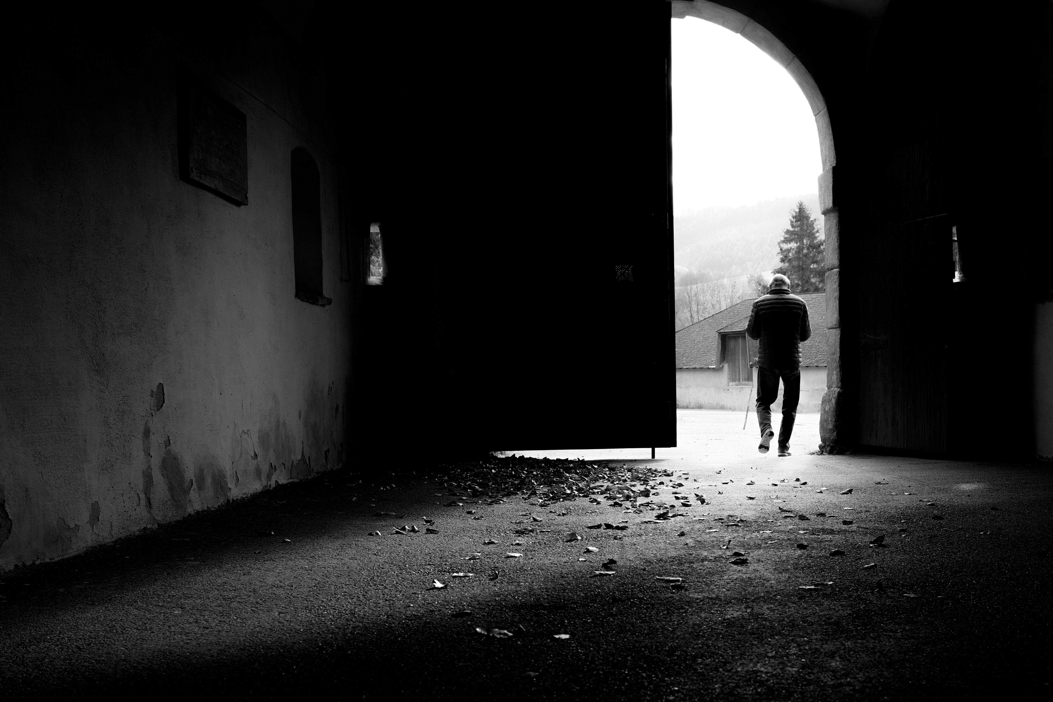 Schwarz-Weiß-Foto von einem 
überdachten Durchgang zu einem hellen Bogenausgang, der linke Türteil ist geschlossen. Auf den Boden liegen verstreuten Blättern. Durch die rechte offene Tür geht eine Person. Durch Tür ist in der Ferne noch ein altes Gebäude, ein hoher einzelner Baum und noch weiter ein Wald zu sehen. 
Im Durchgang sind auf den Seiten Tafeln (nicht erkennbar was drauf steht) und Fenster.