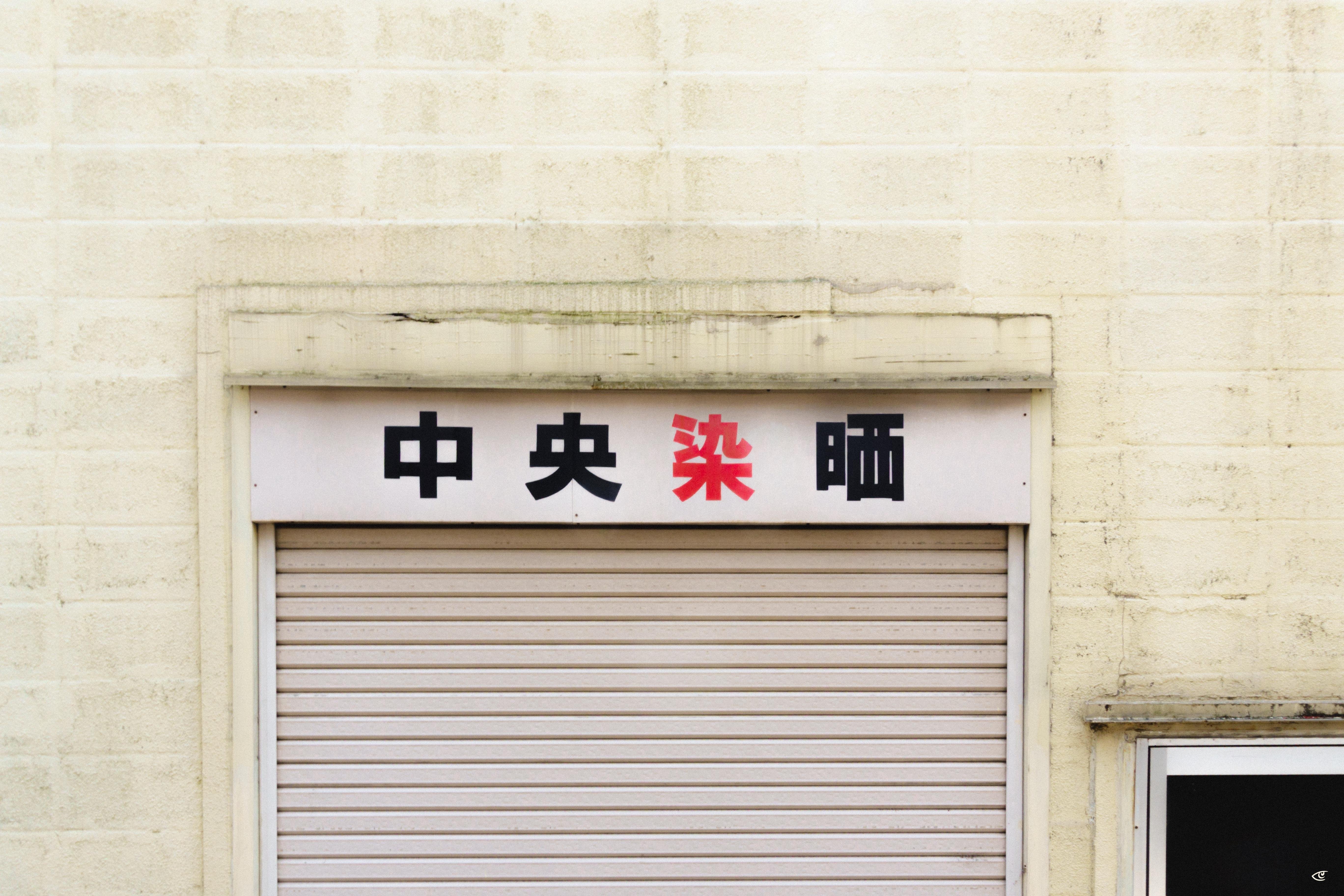 White exterior wall with a closed gray roll-up shutter; above it, a horizontal sign with four Japanese characters in black and red, and a window frame partially visible on the right.