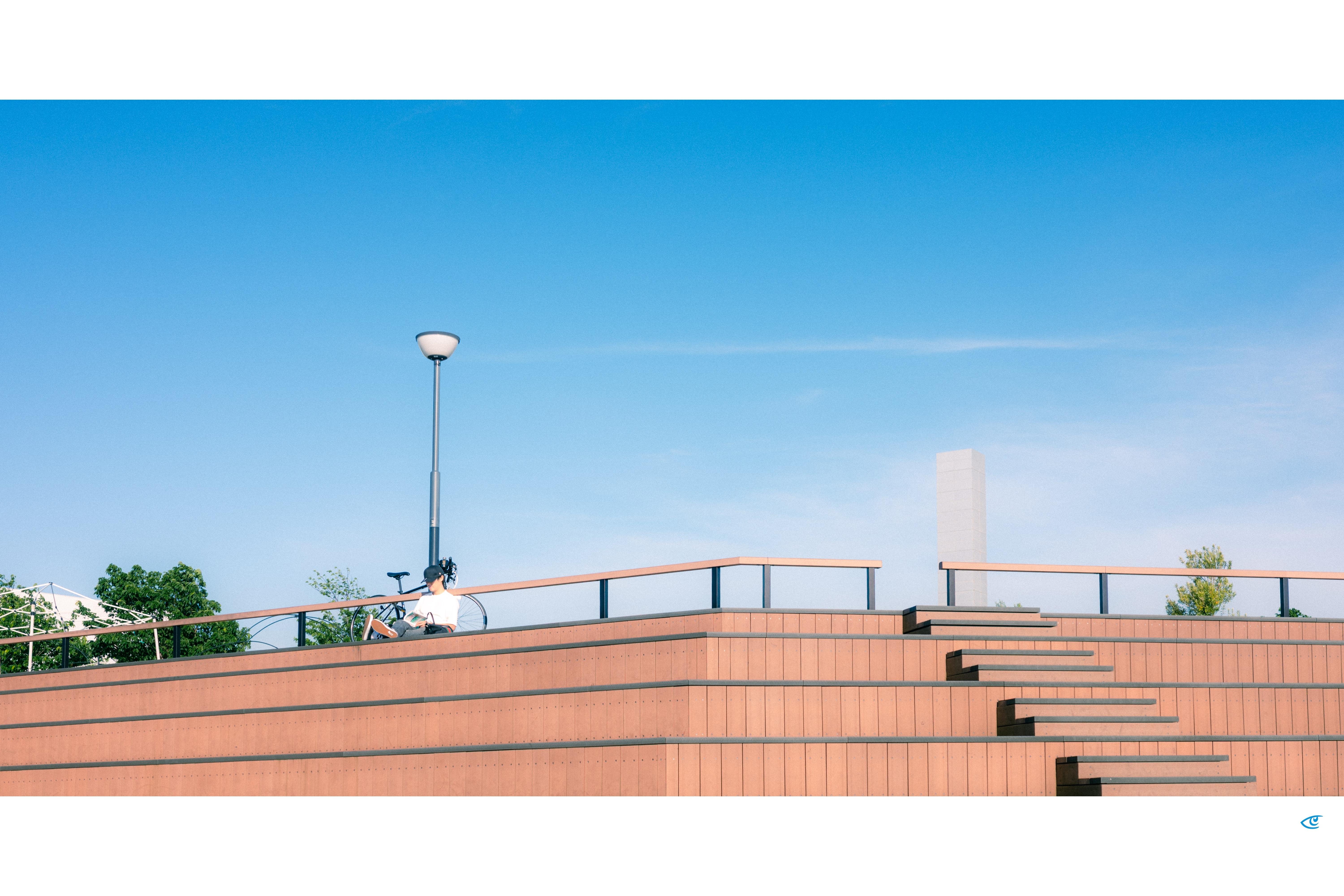 Person sitting beside a bicycle on a terraced brick structure with a railing and a tall streetlamp, under a wide blue sky; a stepped staircase rises on the right with a few trees in the background.
